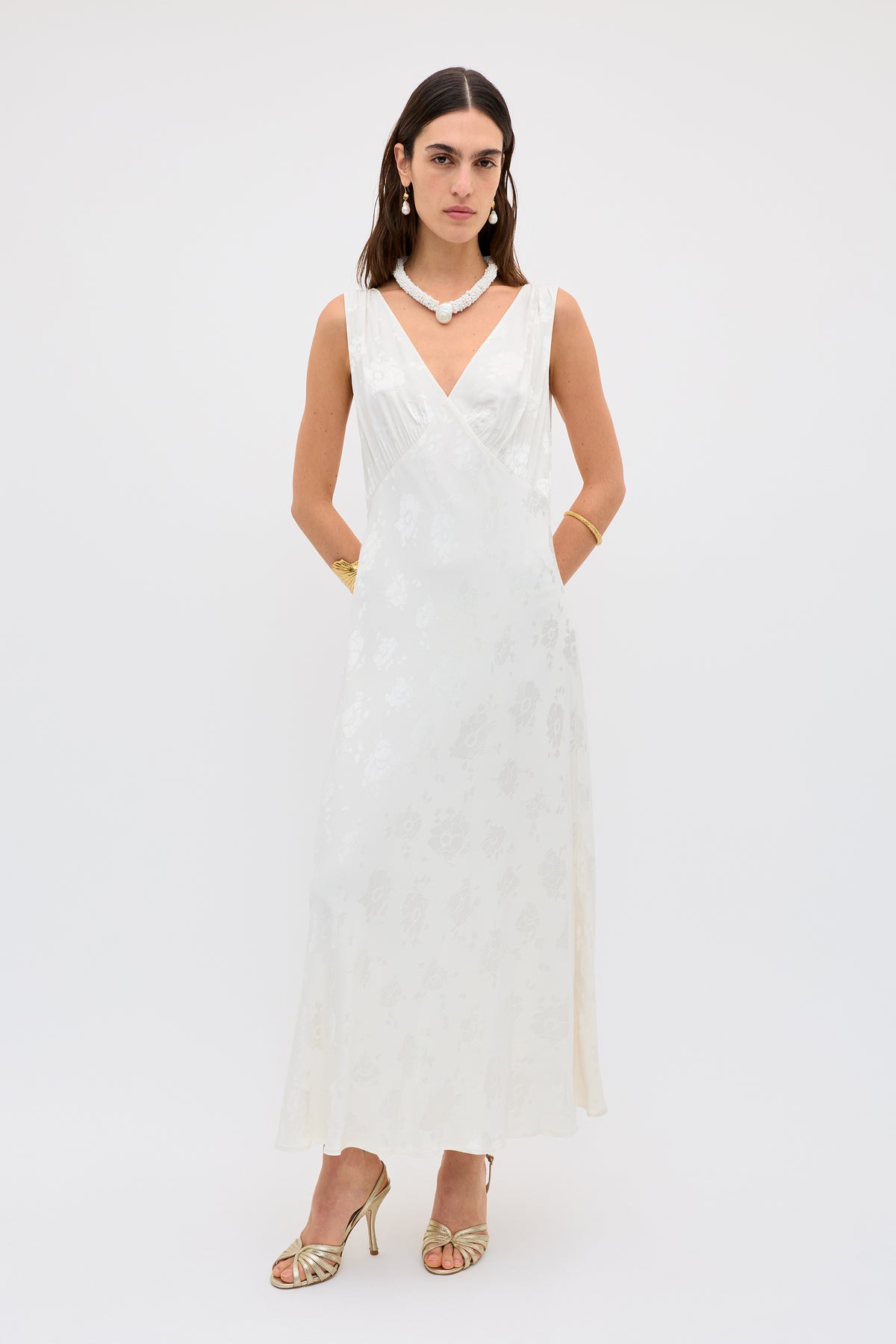 A woman poses against a white background in the Sandrine - Poppy, an ankle-length, sleeveless ivory jacquard dress with a subtle floral pattern, styled with gold high-heeled sandals, jewelry, and a neutral expression.