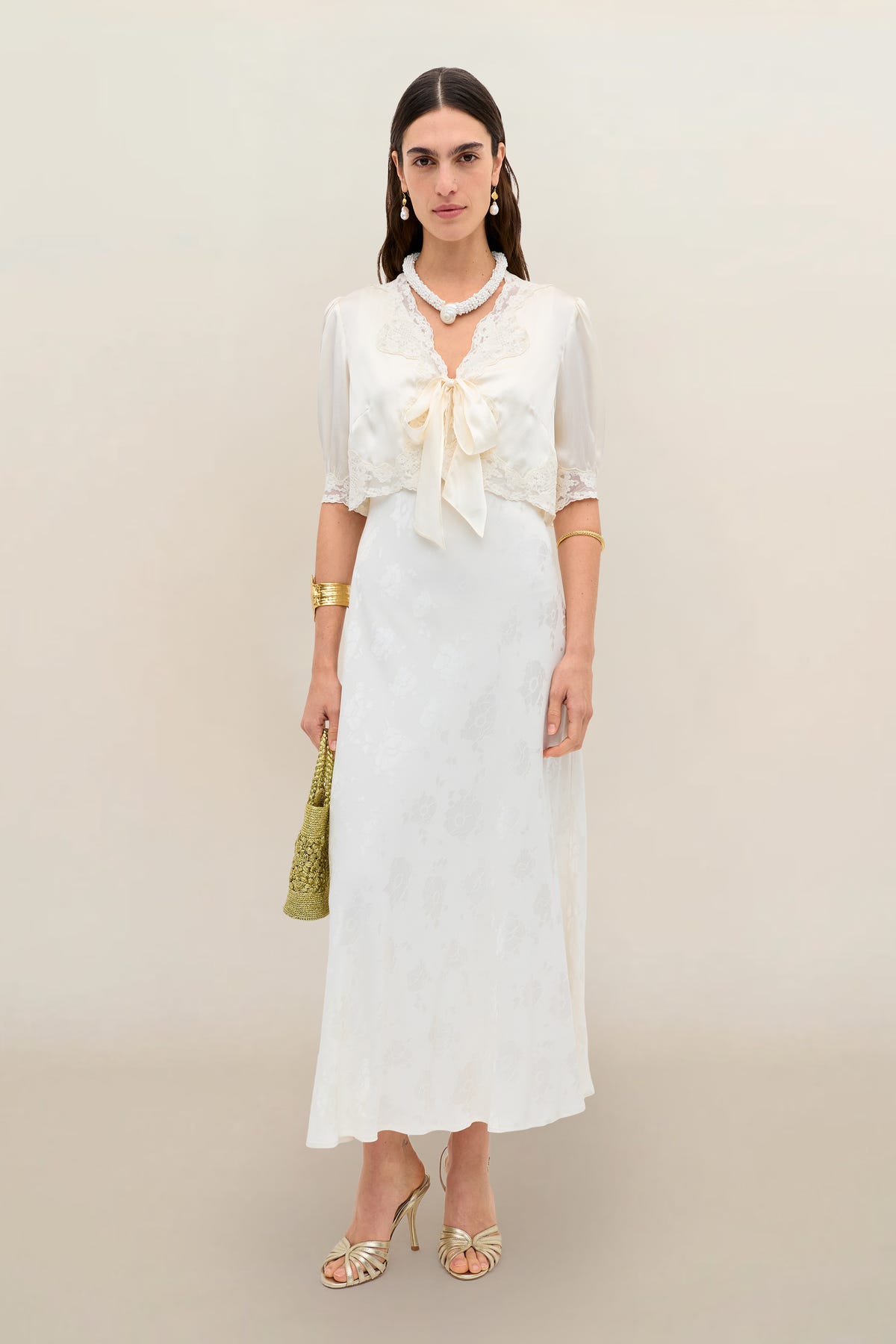 A woman with long dark hair and a neutral expression stands facing forward, wearing the Sandrine - Poppy: a vintage-inspired ivory jacquard calf-length dress with lace trim and a large chest bow, styled with gold heels, bracelets, and a textured gold handbag.