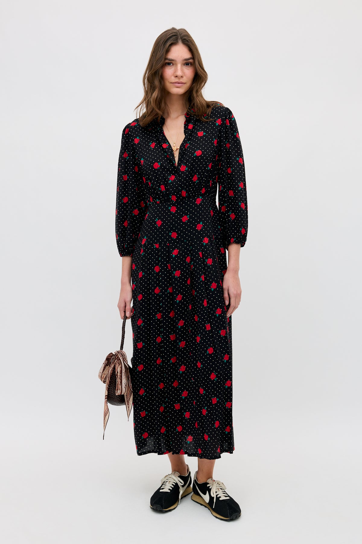 A woman with wavy hair and a relaxed expression stands against a plain background wearing the Izzy - Rose Spot Black long dress with red polka dots, black sneakers, and holding a small striped drawstring bag.
