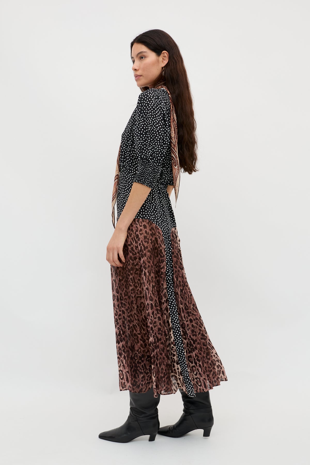 A woman with long brown hair stands sideways wearing the "meg - leopard polka dot" dress—a crepe, round-neck style with black and white polka dots, paired with a brown leopard-print midi skirt and black ankle boots against a plain white background.