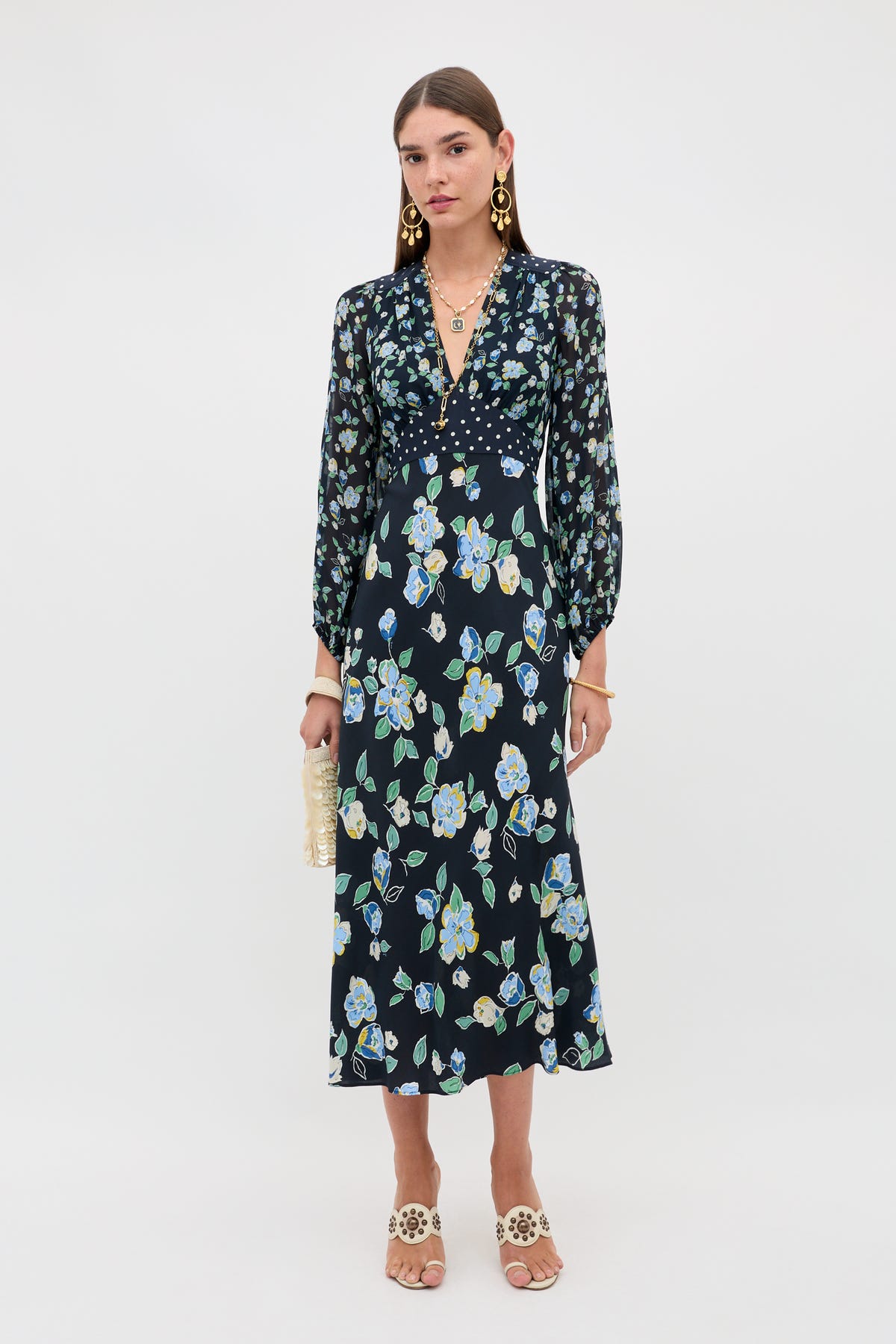 A woman stands against a plain white background wearing the Kaila - Layla Mix Navy long-sleeve floral midi dress, gold statement earrings, beige heeled sandals, and holding a beaded clutch.