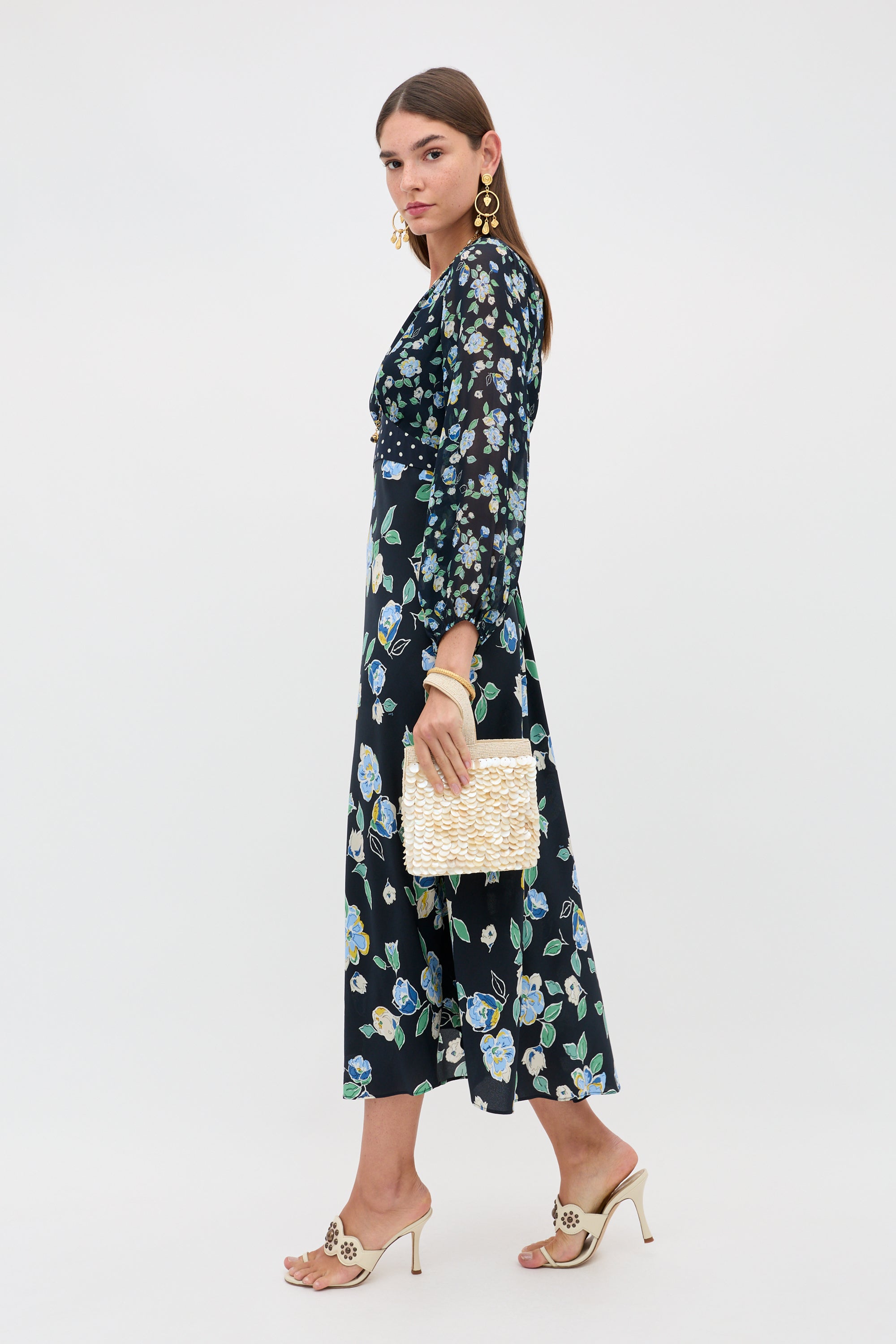 A woman wears the Kaila - Layla Mix Navy floral, long-sleeved midi dress with a woven clutch, beige heeled sandals, and large gold earrings, standing against a plain light background.