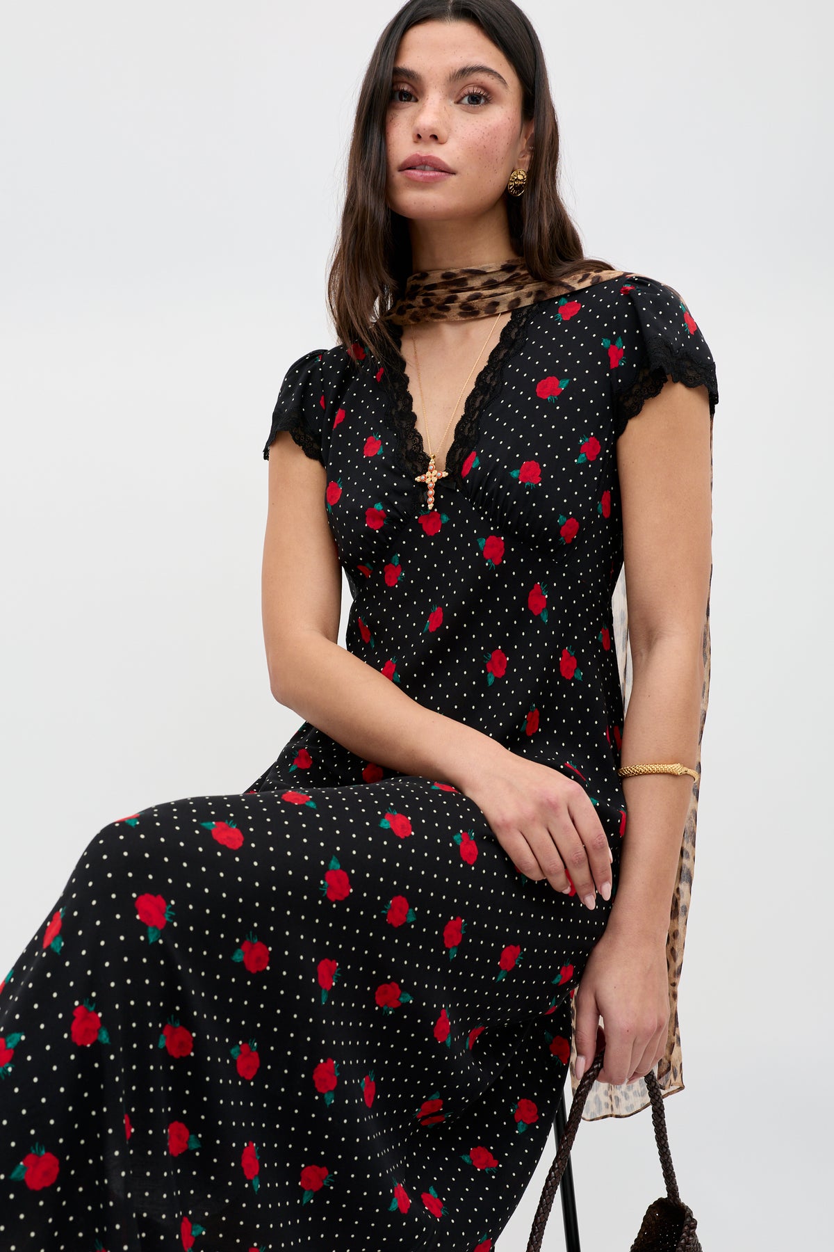 A woman wears the Clarice - Rose Spot Black dress featuring red floral and white polka dot patterns, paired with a brown patterned scarf and a black bag, as she sits with one hand resting on her knee.