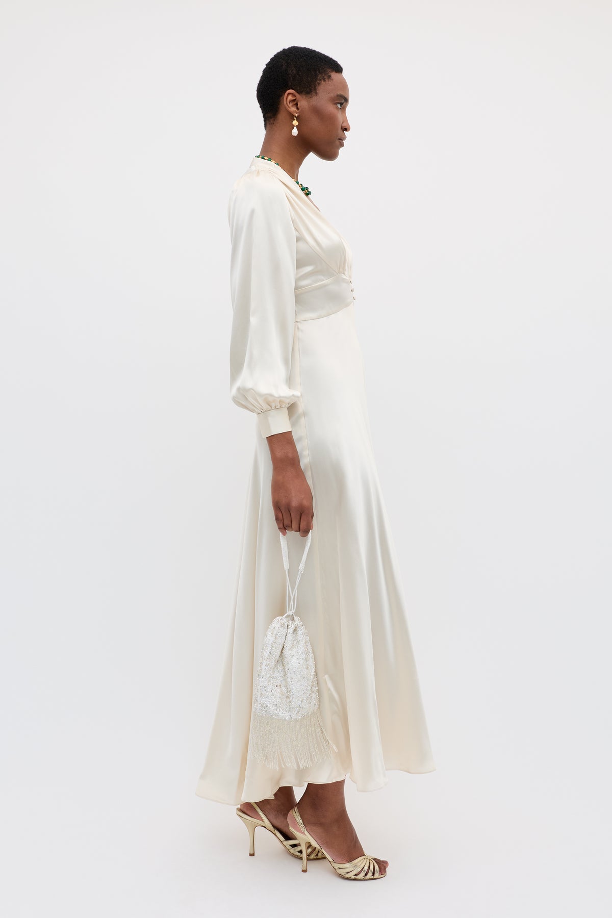 Wearing the Emory - Pearl dress, a woman in cream silk stands in profile with heeled sandals, a small beaded handbag, and drop earrings. She has short hair and poses against a plain white background.