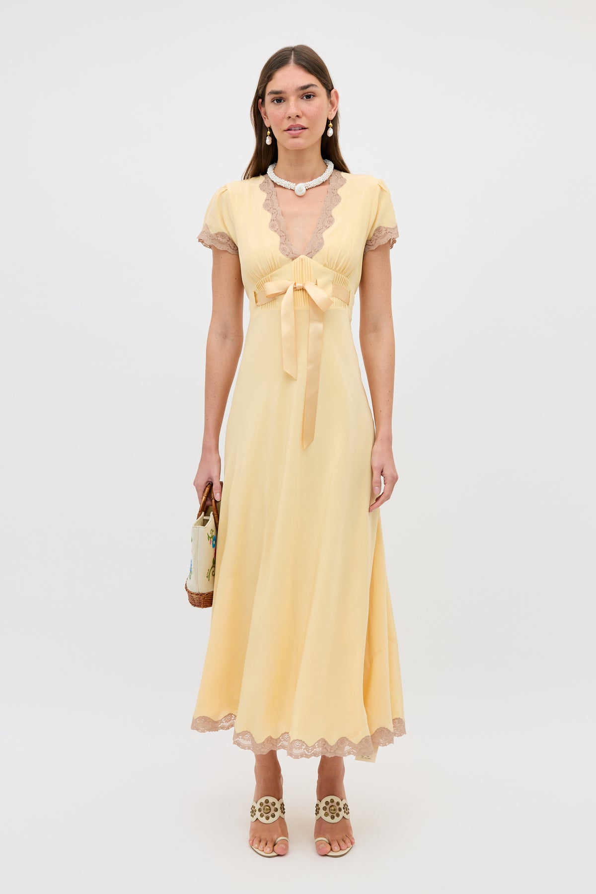 A woman stands against a plain white background wearing the Marlice - Butter Yellow, a long gown with lace trims, satin waist bow, and short sleeves. She holds a small handbag and accessorizes with sandals, pearl earrings, and a necklace.