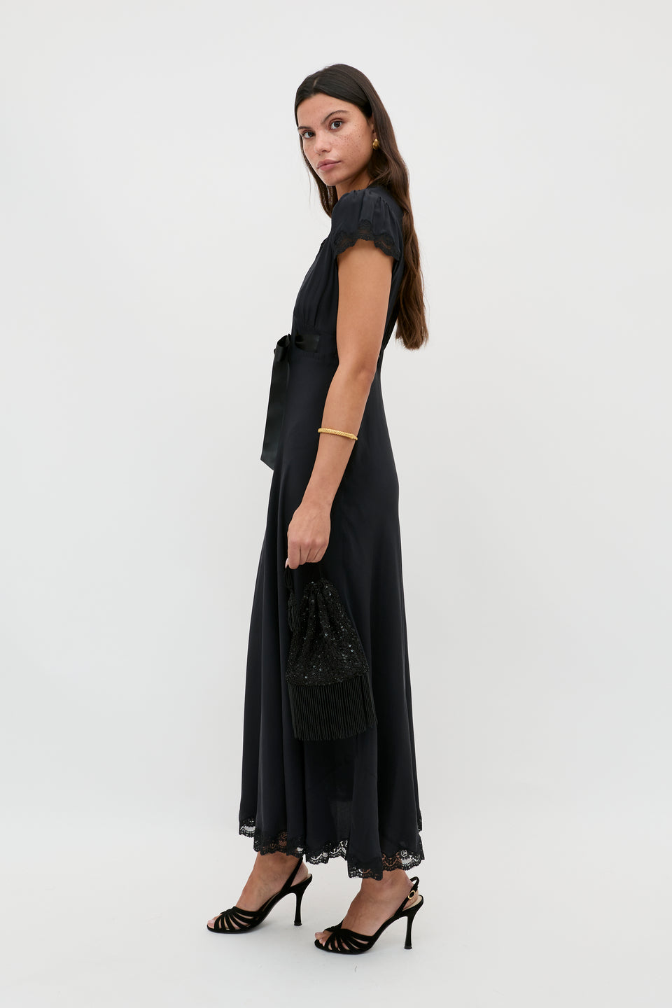 A woman with long brown hair wears the Marlice - Black ankle-length dress with lace trim and a satin bow, styled with black heeled sandals and a matching handbag. She stands sideways against a plain white background.