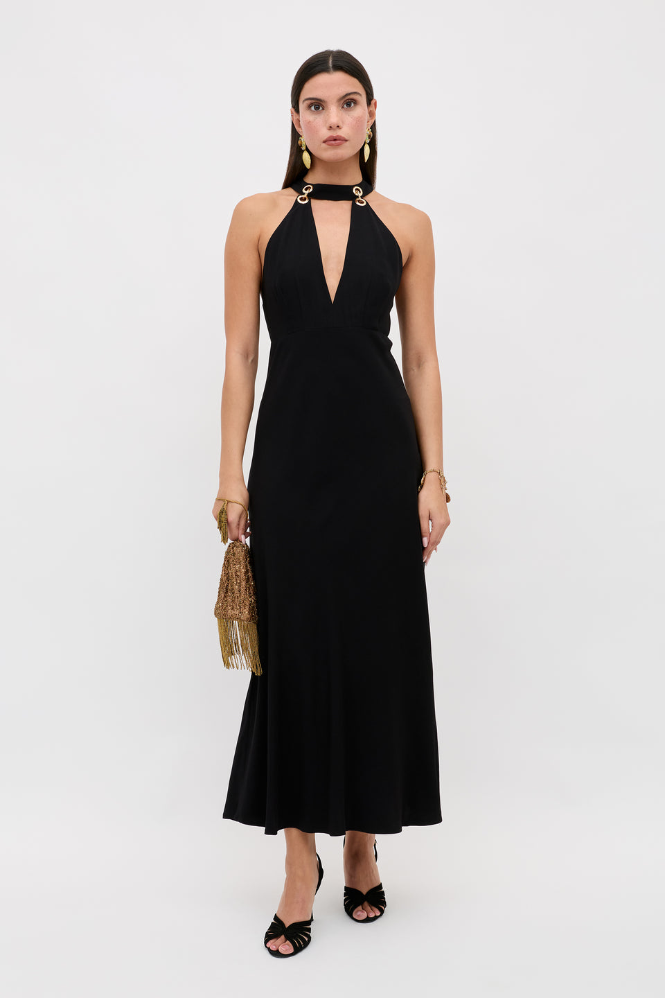 A woman poses against a white background wearing the Eleanora - Black sleeveless, ankle-length dress featuring a deep V neckline and gold ring shoulder details, accessorized with a gold fringe handbag and black open-toe heels.