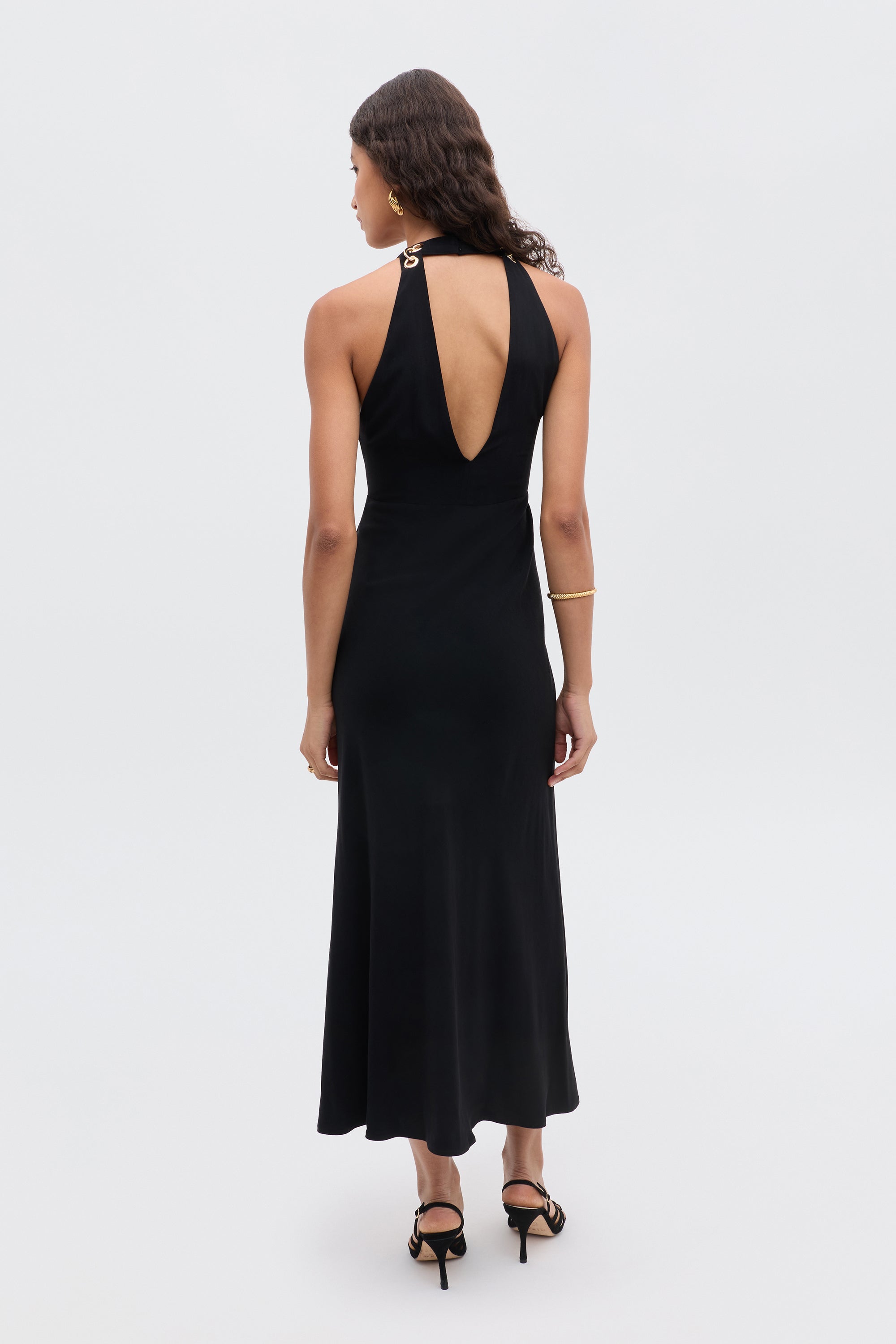 A woman with wavy hair and a gold bracelet stands facing away in the Eleanora - Black sleeveless halter-neck dress with a keyhole back and black heeled sandals, set against a plain light background.