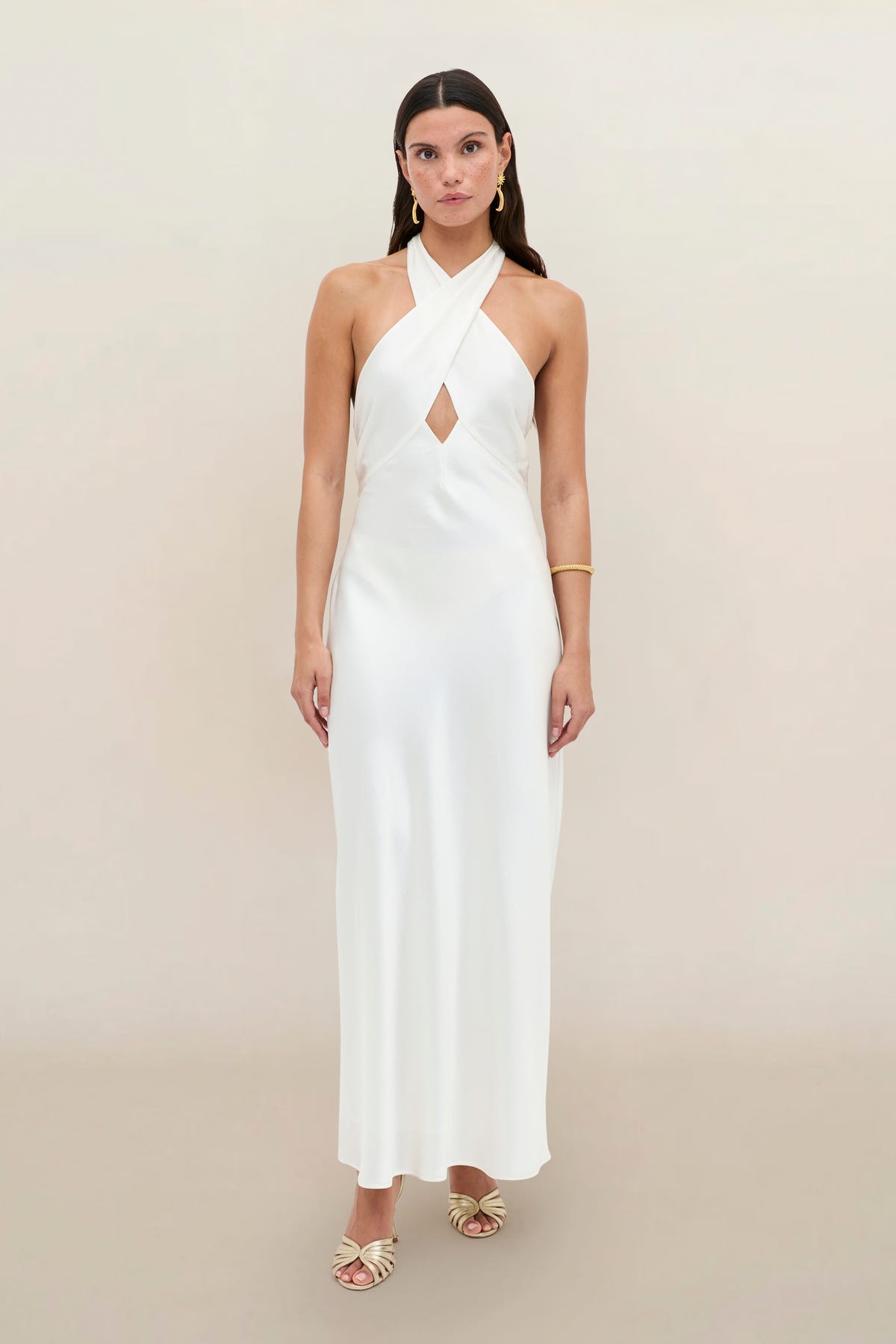 A woman in the Dune Satin Gown—a sleeveless, floor-length white halterneck dress featuring a front keyhole detail—paired with gold sandals and a bracelet, stands against a plain background.