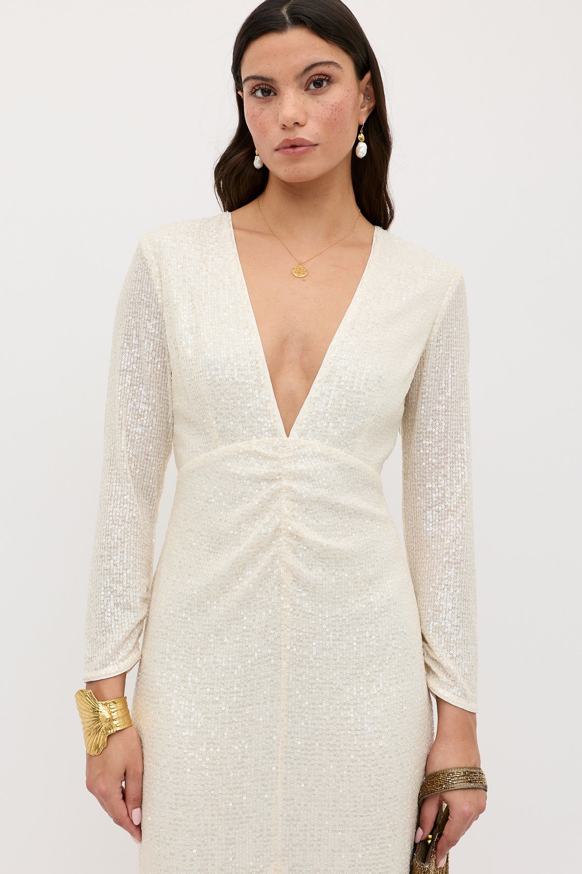 A woman wears the Lorenzo - Pearl: a shimmering ivory dress with long sleeves and a deep V-neckline. She accessorizes with gold earrings, a pendant necklace, gold bracelets, and carries a small clutch bag.