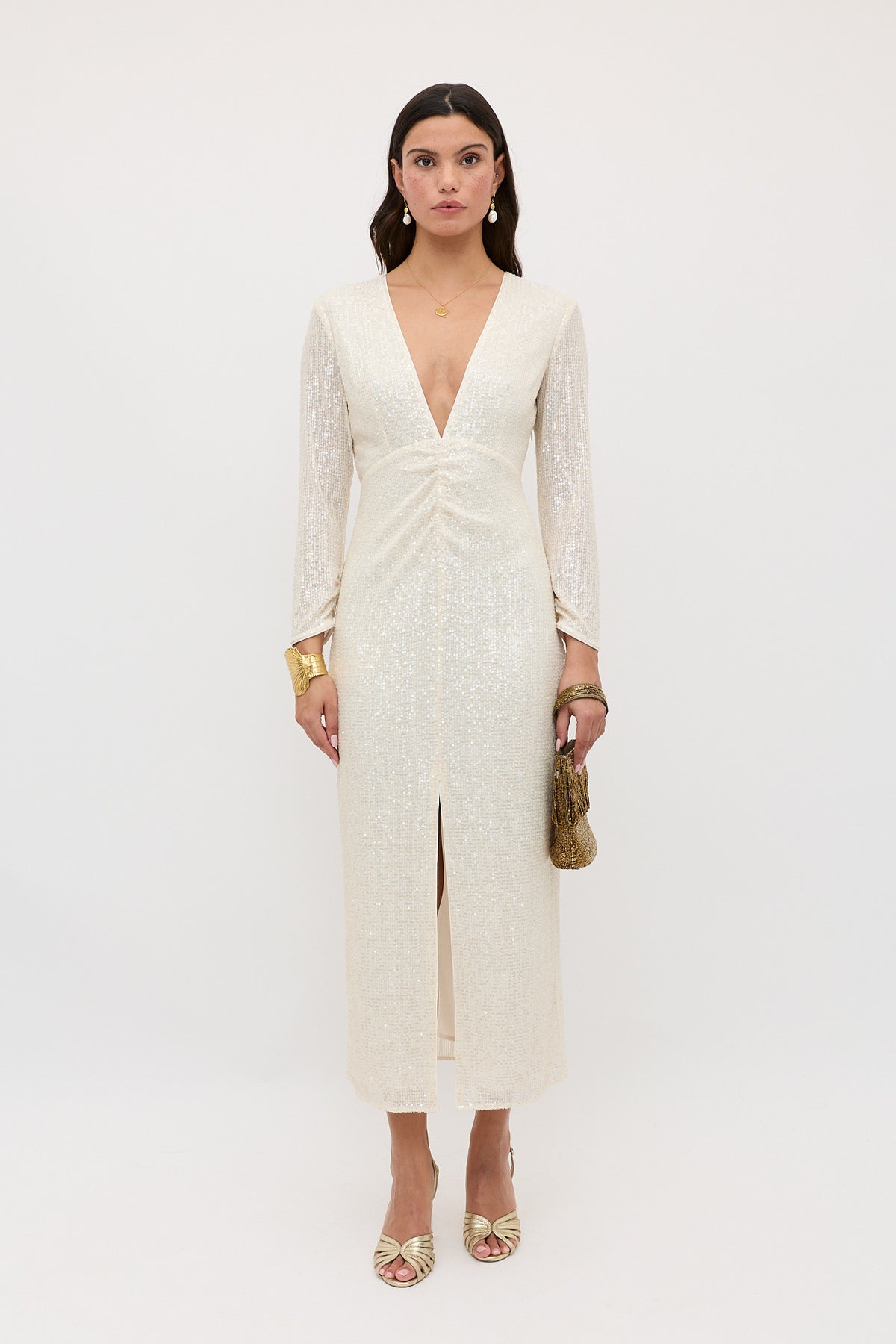 A woman faces forward wearing the Lorenzo - Pearl dress, a long-sleeved white sequin gown with a deep V-neck and front slit. She holds a small gold bag, wears gold sandals and jewelry, all set against a plain white background.