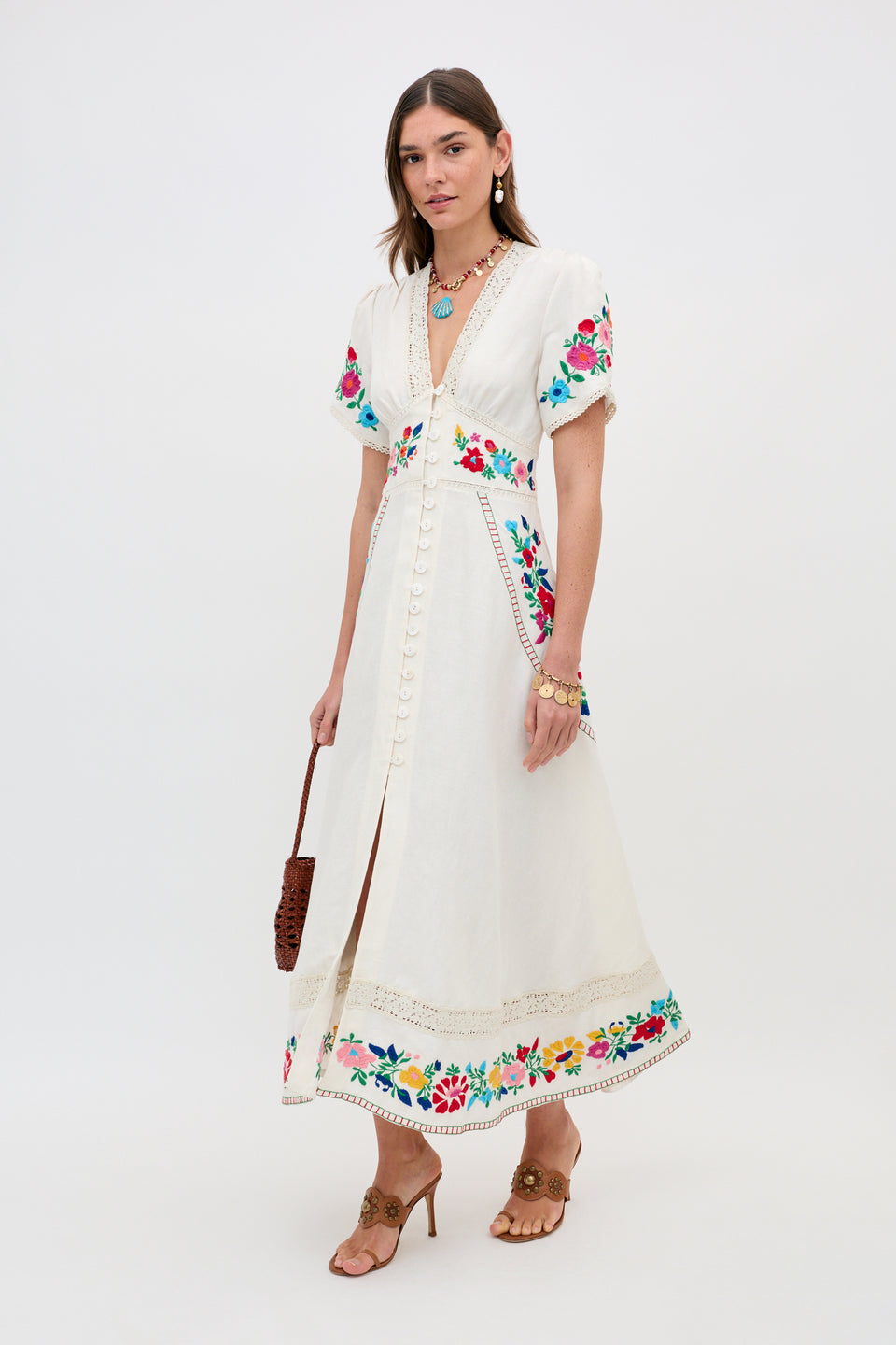 A woman wears the Novara - Anemone Embroidery Cream dress, featuring colorful floral embroidery. She accessorizes with a brown handbag, tan heeled sandals, layered necklaces, and earrings, posing against a plain white background.