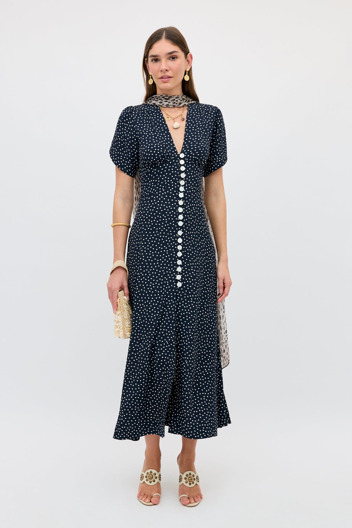 A woman stands against a plain background wearing the Novara - Painted Spot Midnight Navy dress, paired with beige sandals, gold jewelry, a neck scarf, and holding a small woven clutch.