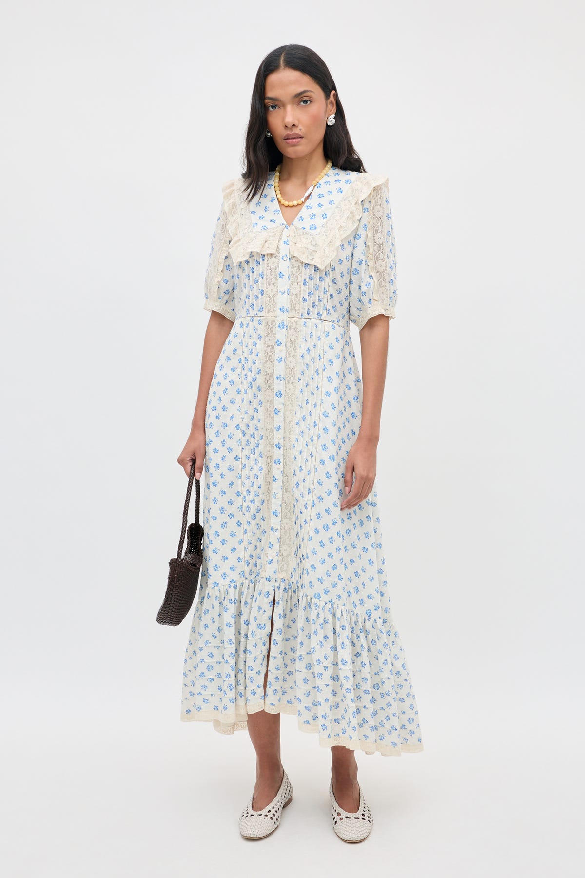 A woman stands against a white background wearing the Edina - Seville Floral Cream dress, which features delicate lace trim. She holds a small brown bag, pairs the outfit with white shoes, and completes her look with long dark hair, earrings, and a necklace.