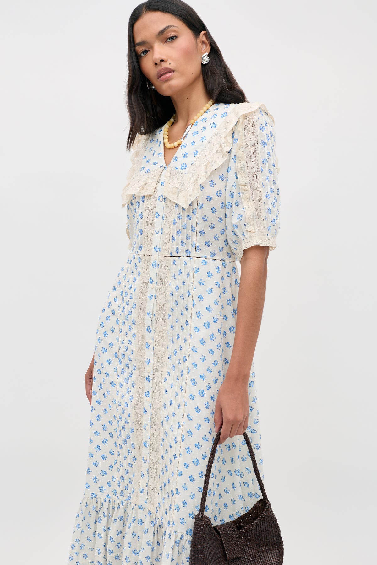 A woman wearing the Edina - Seville Floral Cream dress with lace details, accessorized with pearl earrings, a necklace, and holding a dark woven handbag against a plain background.