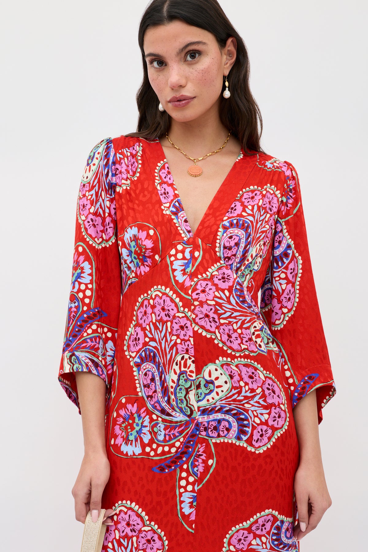 A woman wears the Dahlia - Butterfly Bloom Jacquard Red dress, featuring bold pink, purple, and blue floral patterns, a V-neckline, and three-quarter sleeves. She accessorizes with gold jewelry and a small beige bag.