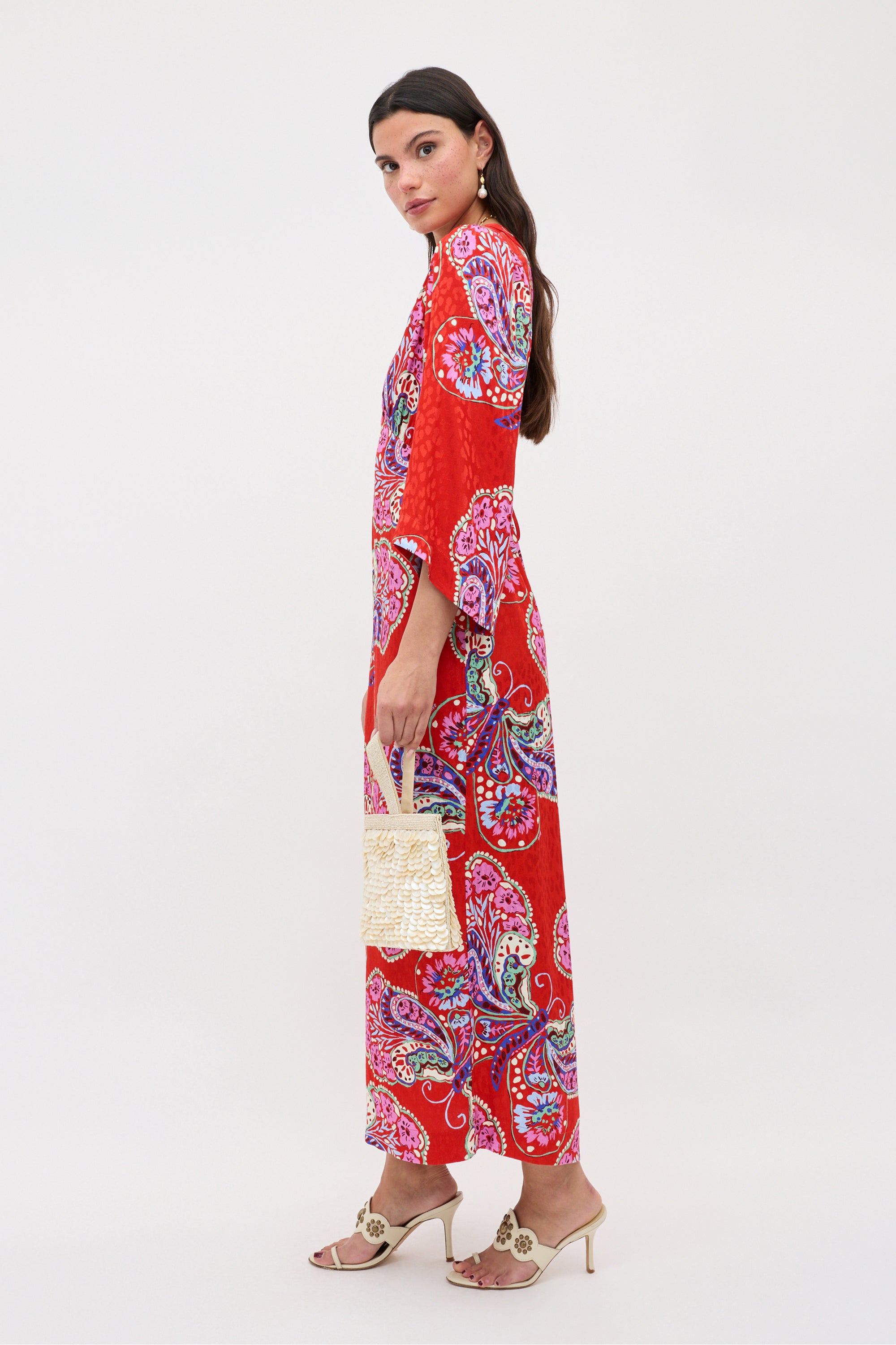 Wearing the Dahlia - Butterfly Bloom Jacquard Red dress, a woman with long dark hair stands sideways, holding a woven handbag and wearing open-toe heeled sandals and neutral earrings against a plain background.