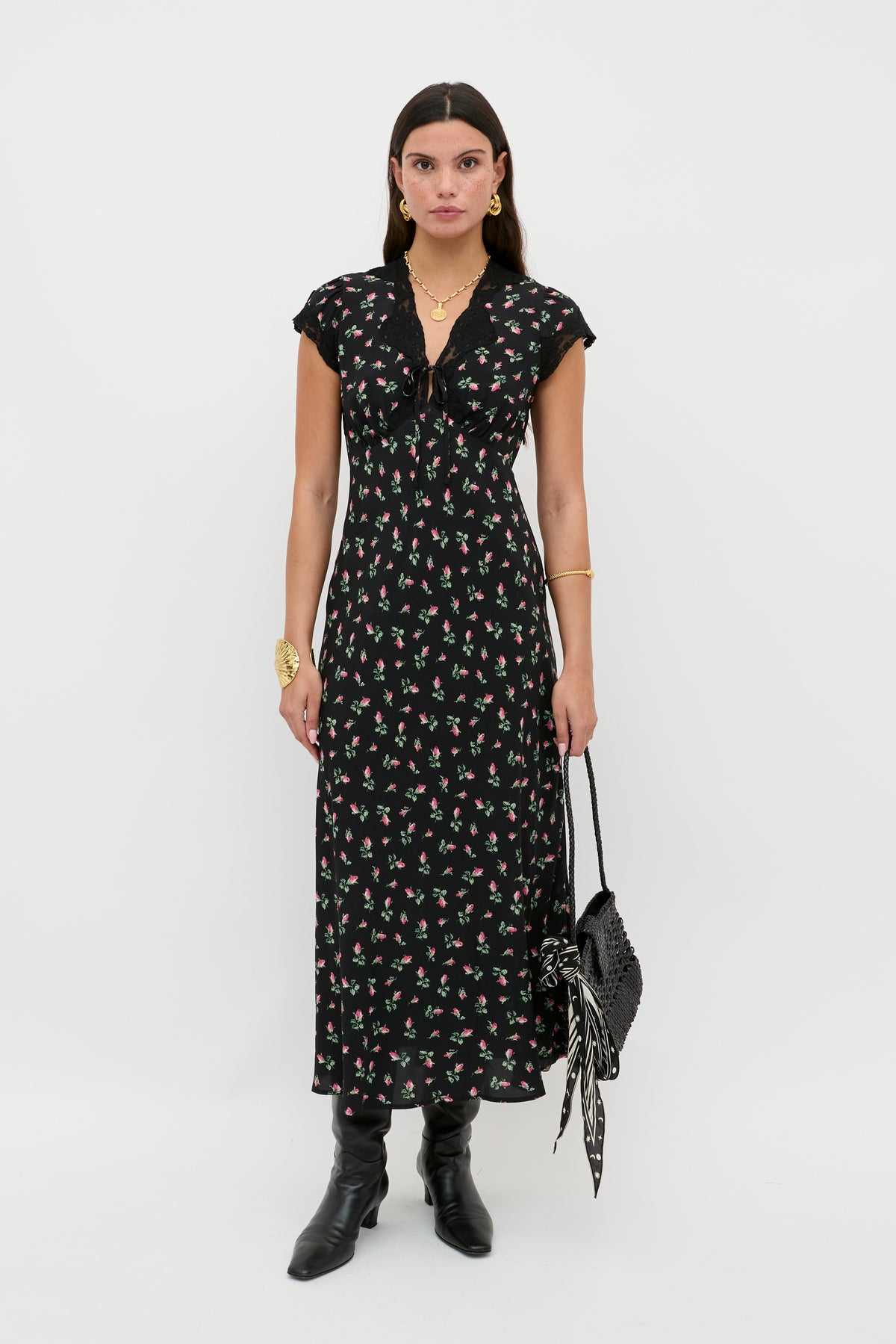 A woman poses against a white background in the Amari - Sienna Bud Black short-sleeved midi dress with a gathered neckline, styled with black boots, gold jewelry, and a black handbag adorned with a patterned scarf.