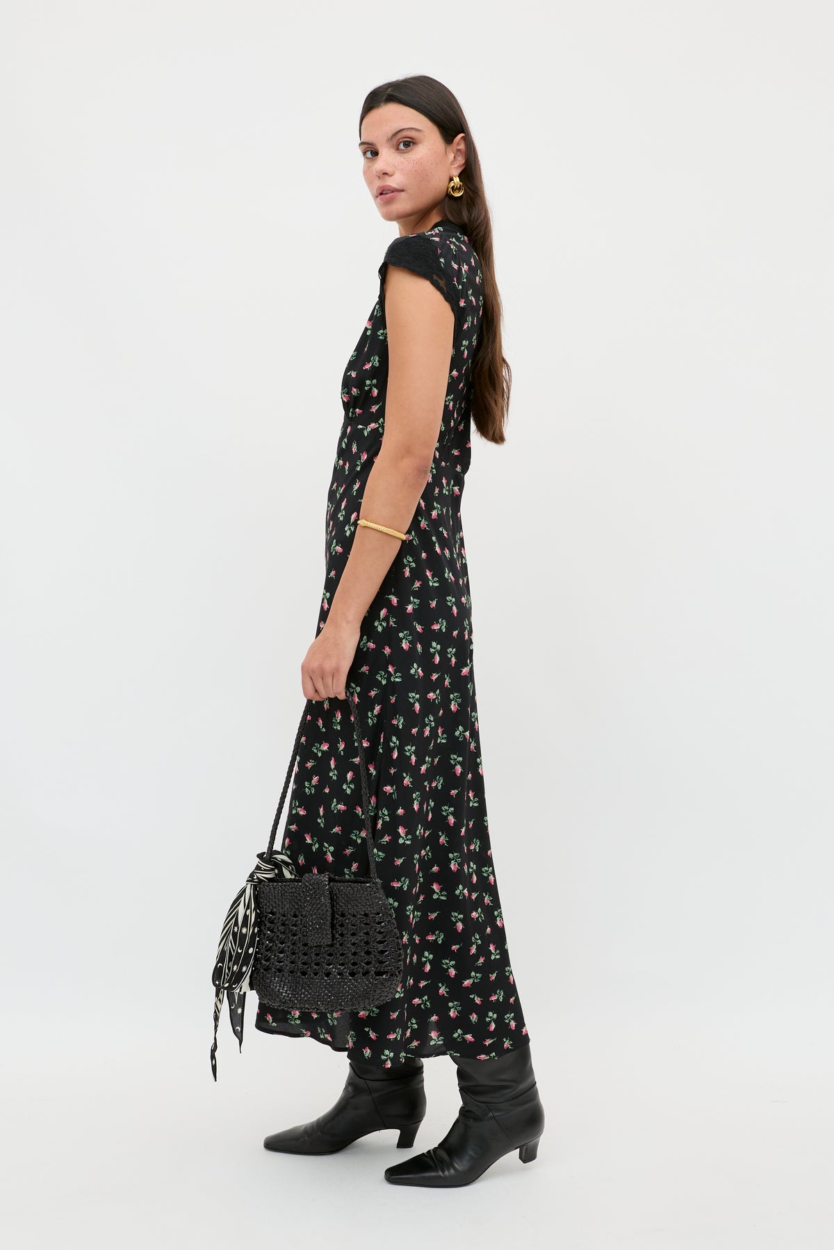 A woman wearing the Amari - Sienna Bud Black dress, black boots, gold jewelry, and holding a black woven handbag stands sideways against a plain white background.