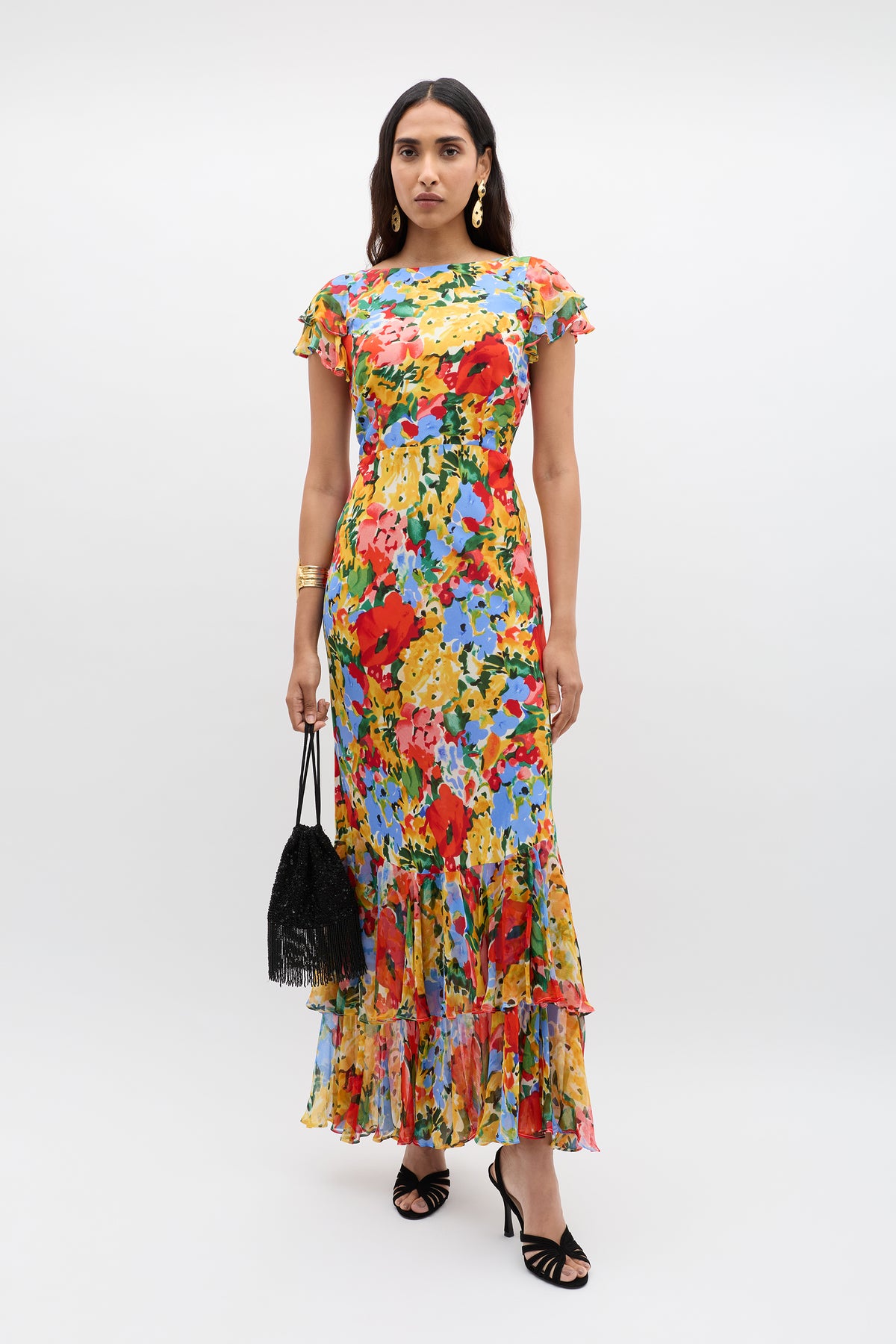 Against a plain background, a woman wears the vintage-inspired Ordinia – Wedding Floral Multi gown—an ankle-length, floral-patterned dress with a ruffled hem. She accessorizes with black heels, hoop earrings, a gold bracelet, and a black handbag.