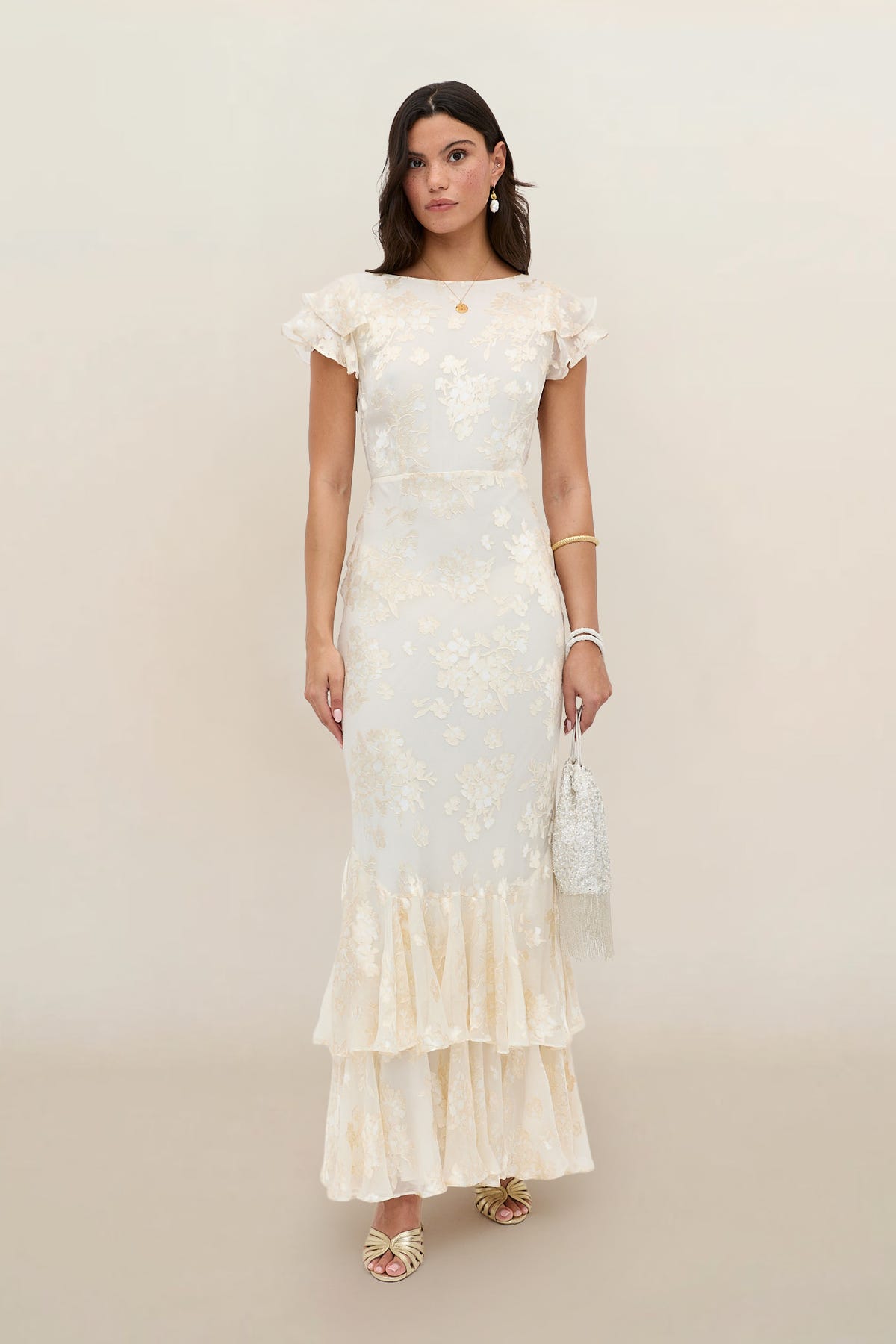 A woman wears the Ordinia - Bouquet Devore Cream gown with a floral print, short ruffled sleeves, and a tiered ruffle hem. She holds a beaded handbag, accessorizes with gold jewelry and heeled sandals, standing against a neutral background.