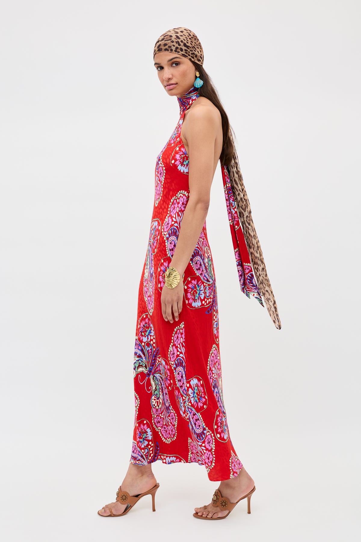 A woman in profile wears the Maura - Butterfly Bloom Jacquard Red long, sleeveless dress, paired with a leopard-print headscarf, gold bracelet, blue earrings, and brown heeled sandals against a plain background.