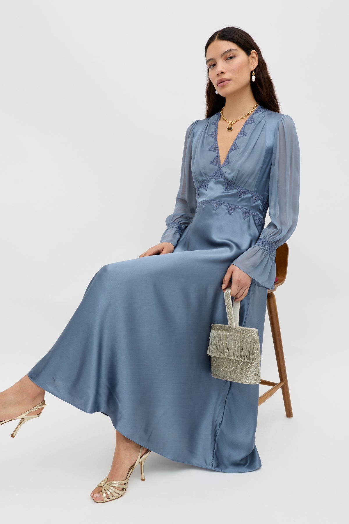 A woman in the Arlena - Stone Blue long-sleeved dress sits on a stool, holding a small fringed handbag and wearing gold strappy heels and jewelry, posing against a plain light background.