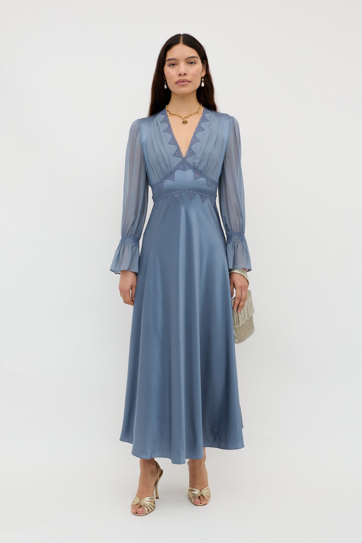 A woman stands against a white background wearing the Arlena - Stone Blue, a long, silky dress with sheer sleeves and a deep V-neckline. She accessorizes with a small textured clutch, gold heels, earrings, and a necklace.