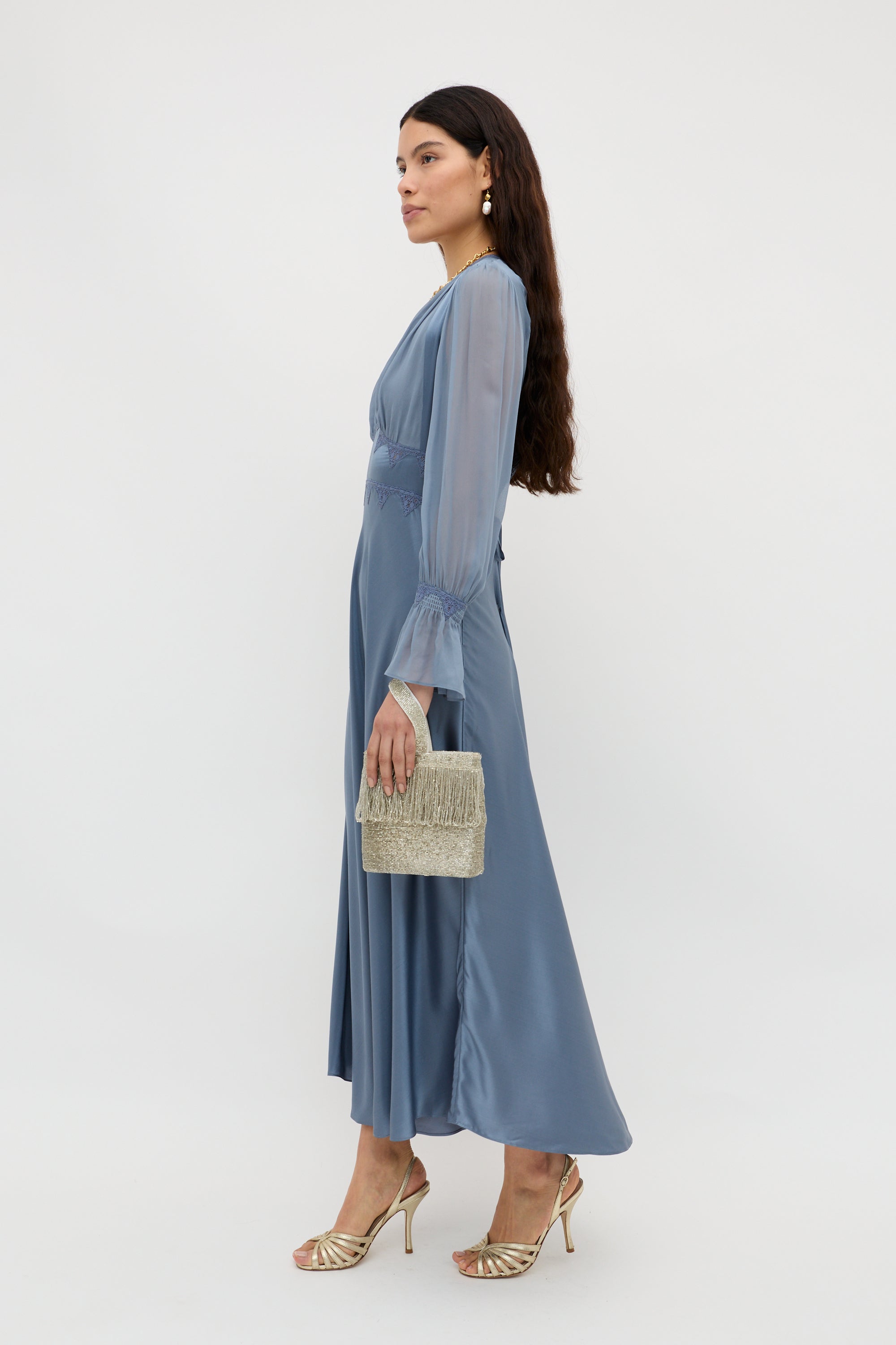A woman with long, wavy brown hair poses in profile against a plain background, wearing the Arlena - Stone Blue long silky dress with gold strappy heels, earrings, and holding a beaded clutch.