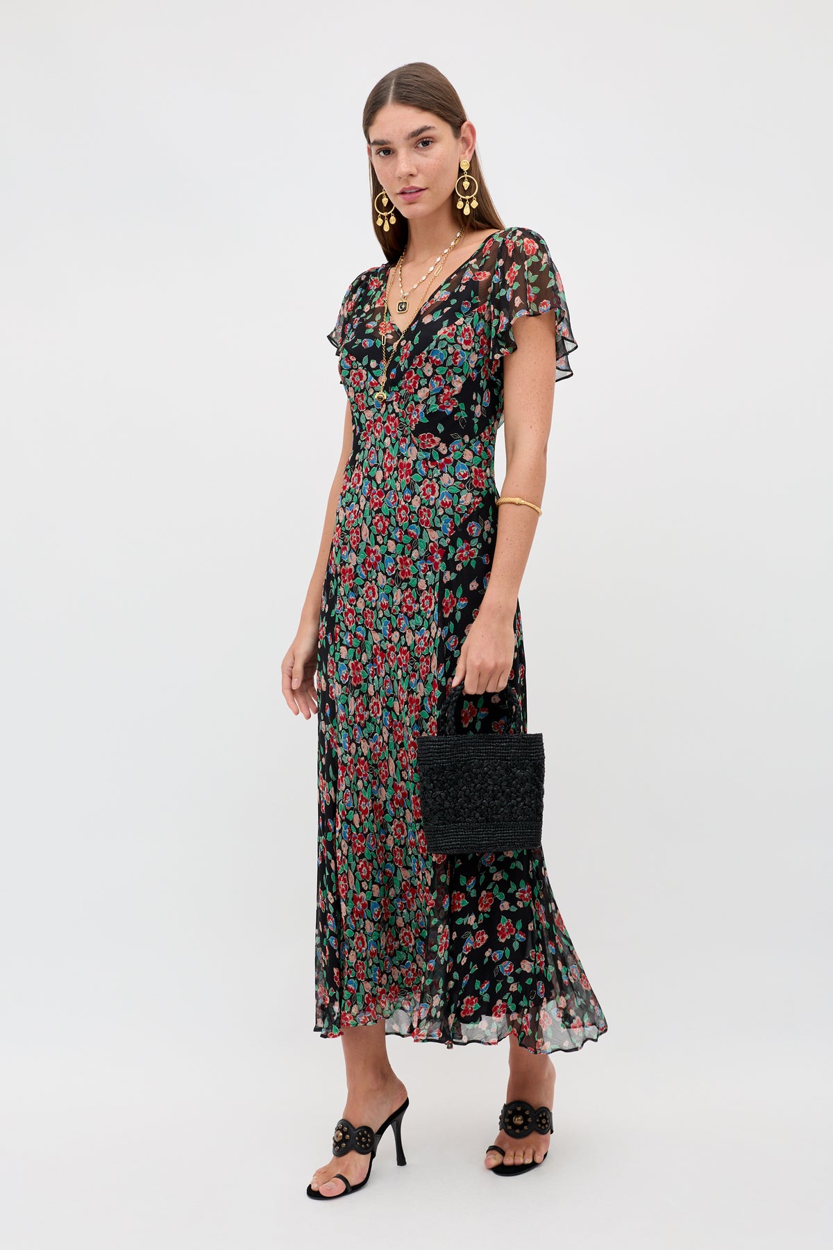 A woman models the Bexley - Jeannie Mix Charcoal, a black sheer floral midi dress with short flutter sleeves, paired with black heeled sandals, gold jewelry, and a textured black handbag against a plain white background.