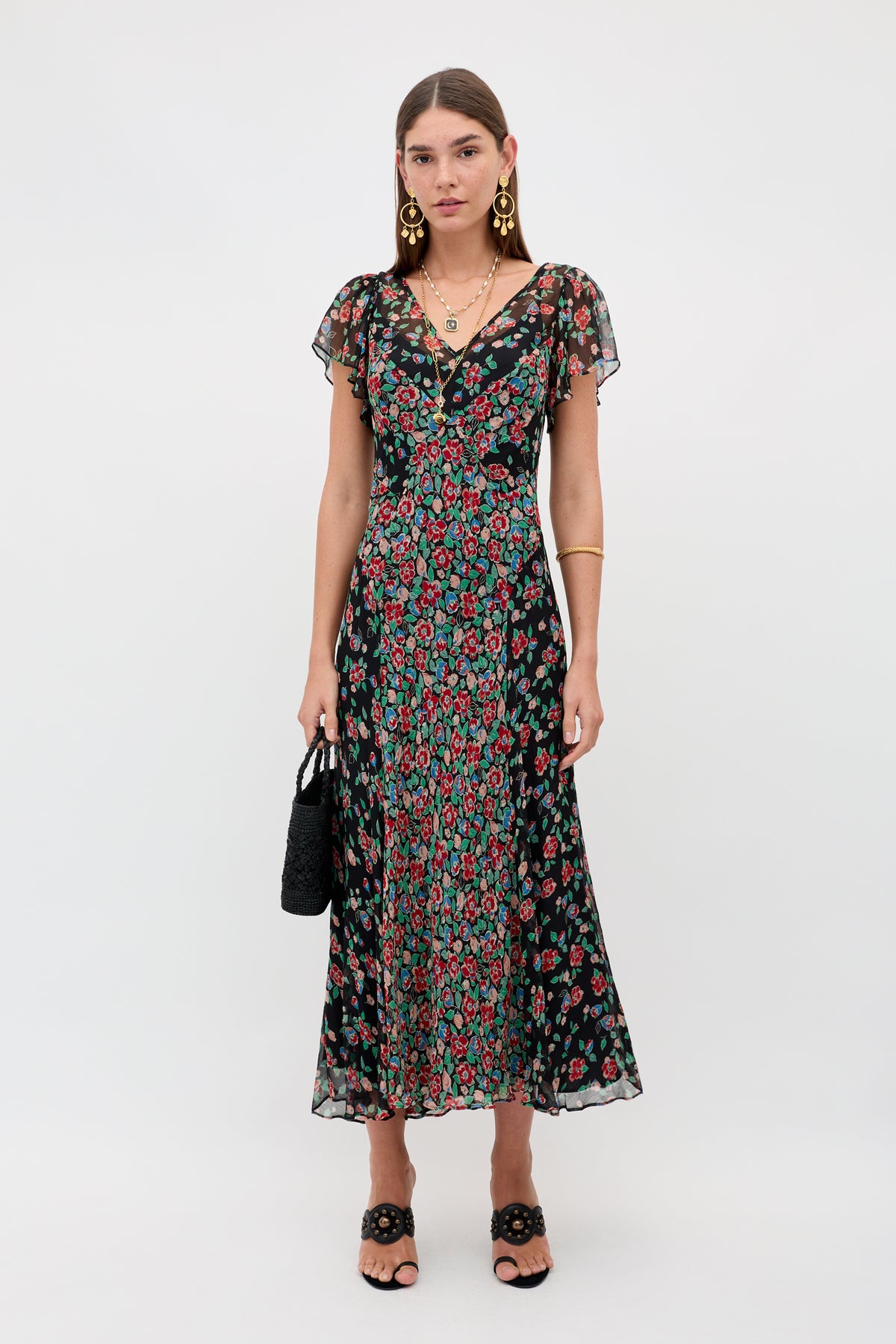A woman stands against a plain background wearing the Bexley - Jeannie Mix Charcoal, an ankle-length black dress with a red and green floral pattern, short flutter sleeves, black sandals, gold jewelry, and holding a black textured handbag.