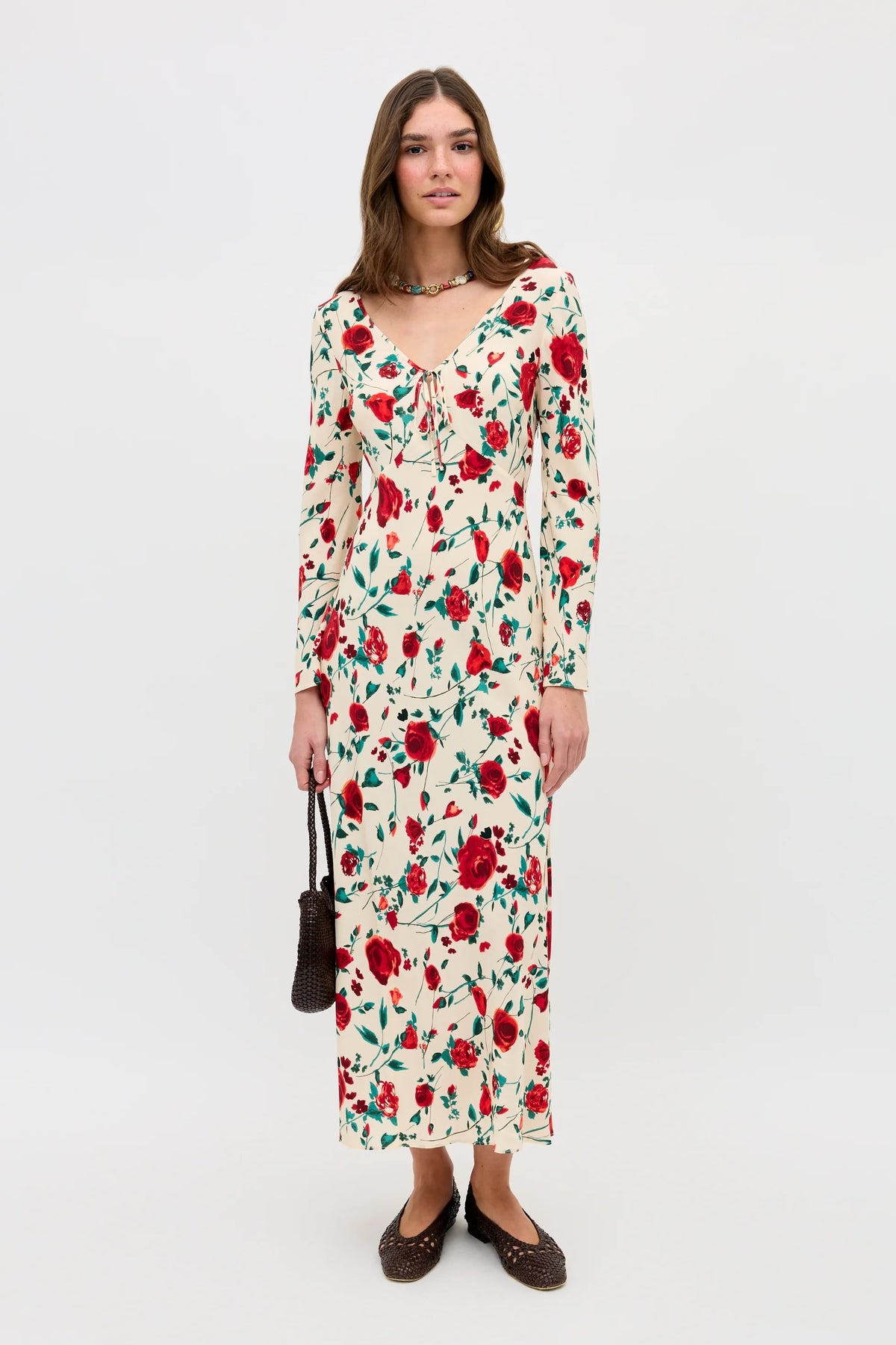 A woman stands against a plain white background wearing the Gwenyth - Claudia Rose Cream dress with red rose and green leaf print, brown flats, a necklace, and holding a small brown handbag.