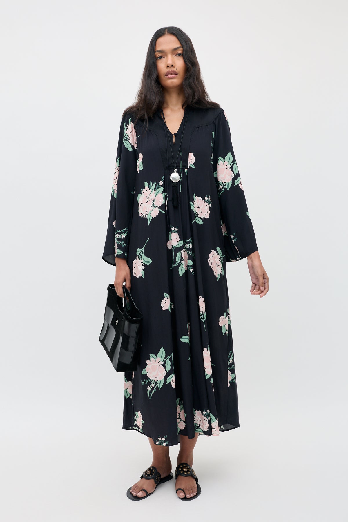 A woman wearing the Lucilla - Peonies Scatter Black dress, featuring pink and green floral patterns, paired with black sandals and a black handbag, stands against a plain light background.