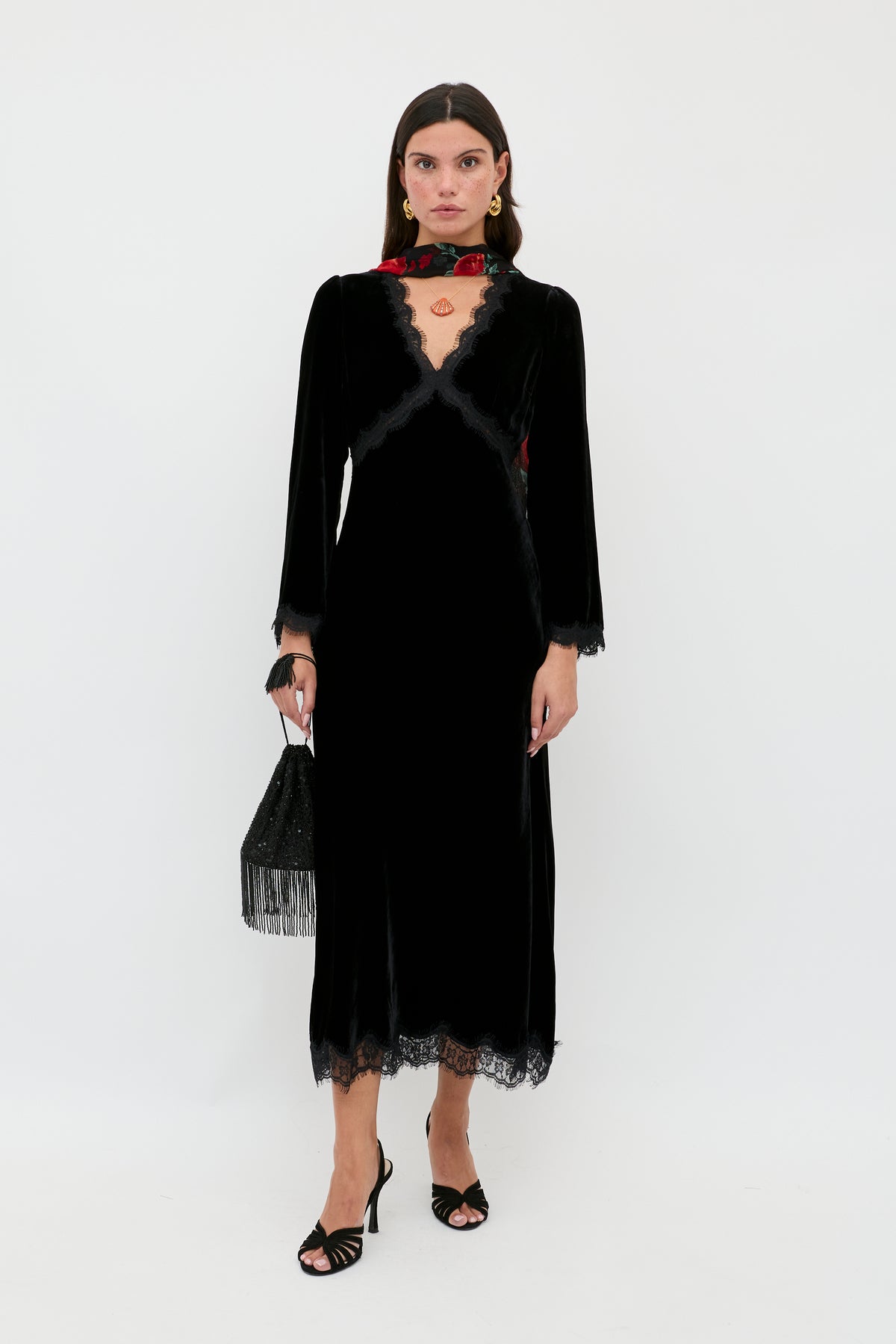 A woman stands against a white background in the Alessi - Black long dress with lace and velvet, holding a black fringed handbag. She pairs it with black open-toe heels, a red floral choker, and wears her hair straight and loose.