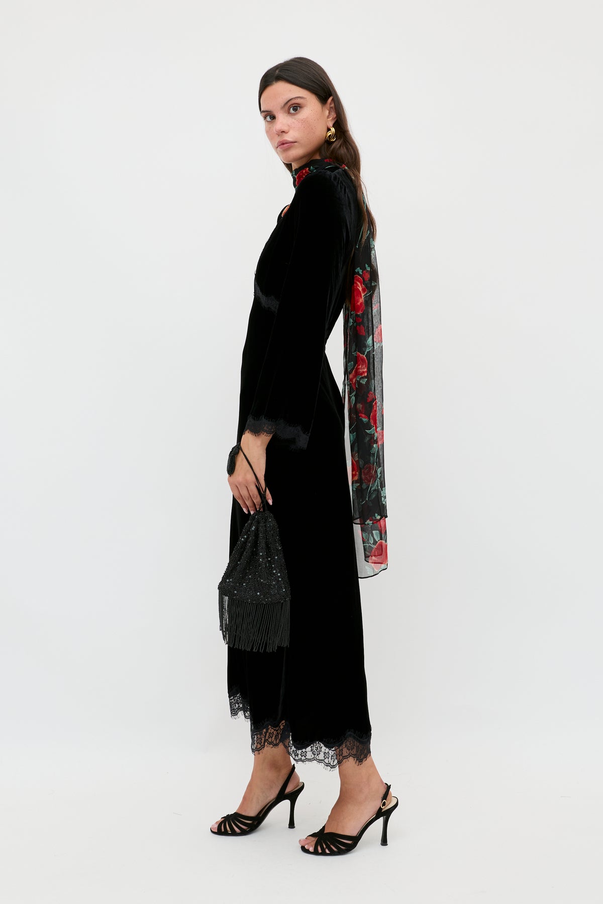 A woman stands sideways against a plain white background in the Alessi - Black luxurious velvet dress with lace detailing, holding a beaded black handbag, wearing black strappy heels, gold earrings, and a floral scarf over one shoulder.