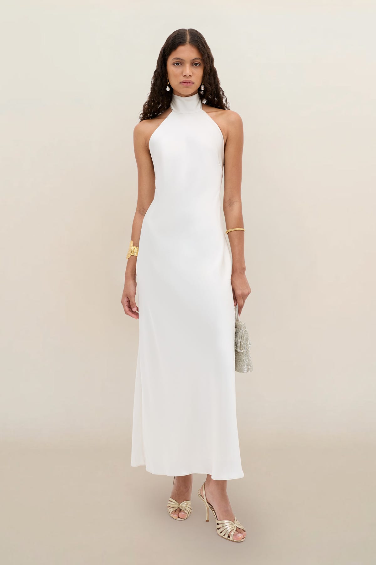A woman wearing the Arista - Pearl sleeveless, high-neck, floor-length white dress with open shoulders, paired with gold strappy heels, gold jewelry, and a small handbag, stands against a plain beige background.