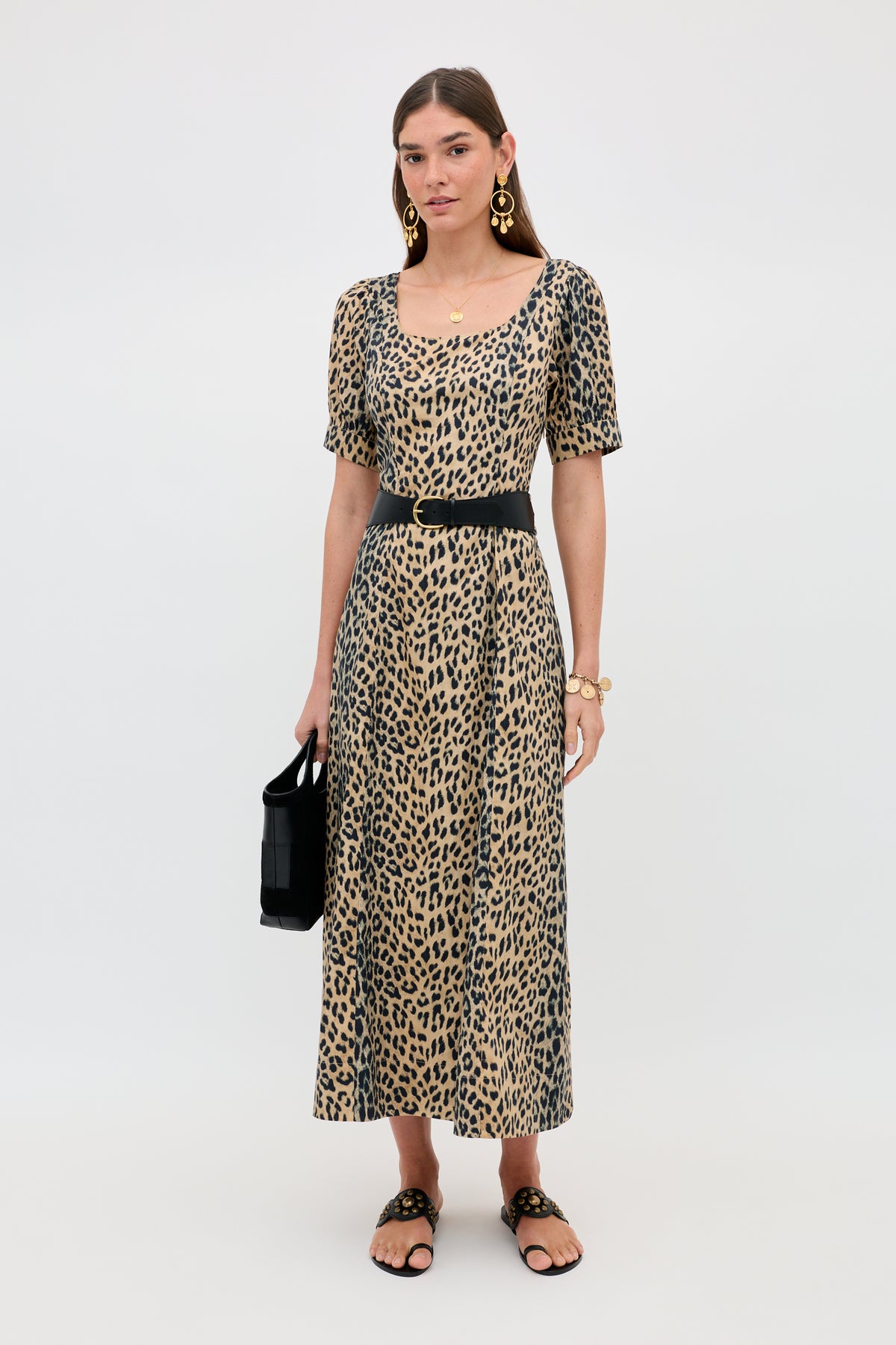 A woman with straight brown hair and a neutral expression stands against a plain background, wearing the Kathleen - Nina Leopard Brown dress, paired with a black belt, black sandals, gold statement earrings, and holding a black handbag.