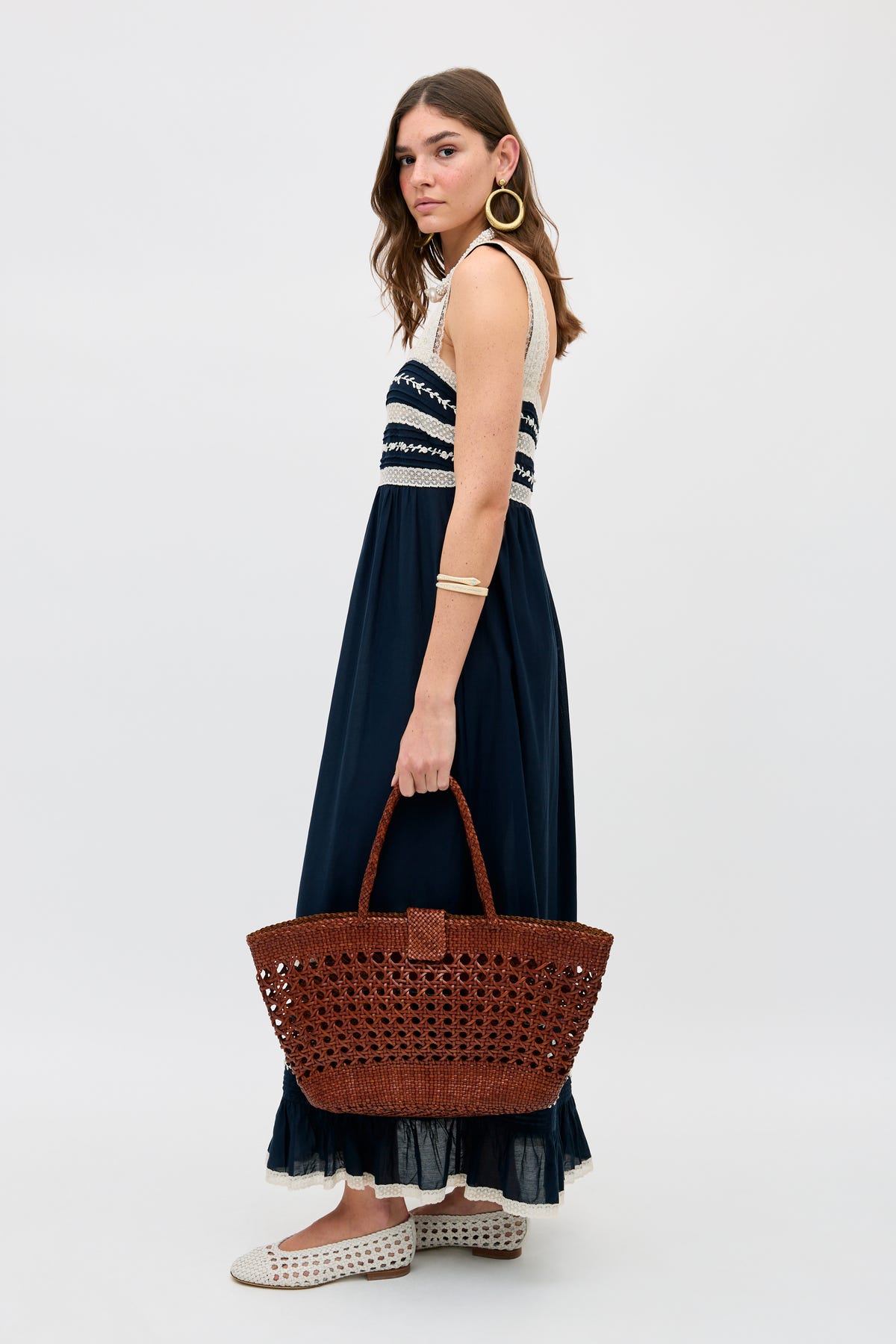 A woman with long brown hair in a sleeveless navy and white maxi dress holds the Lucine - Tan bag, crafted from woven leather. She wears gold hoop earrings, a gold bracelet, and white woven shoes against a plain light background.