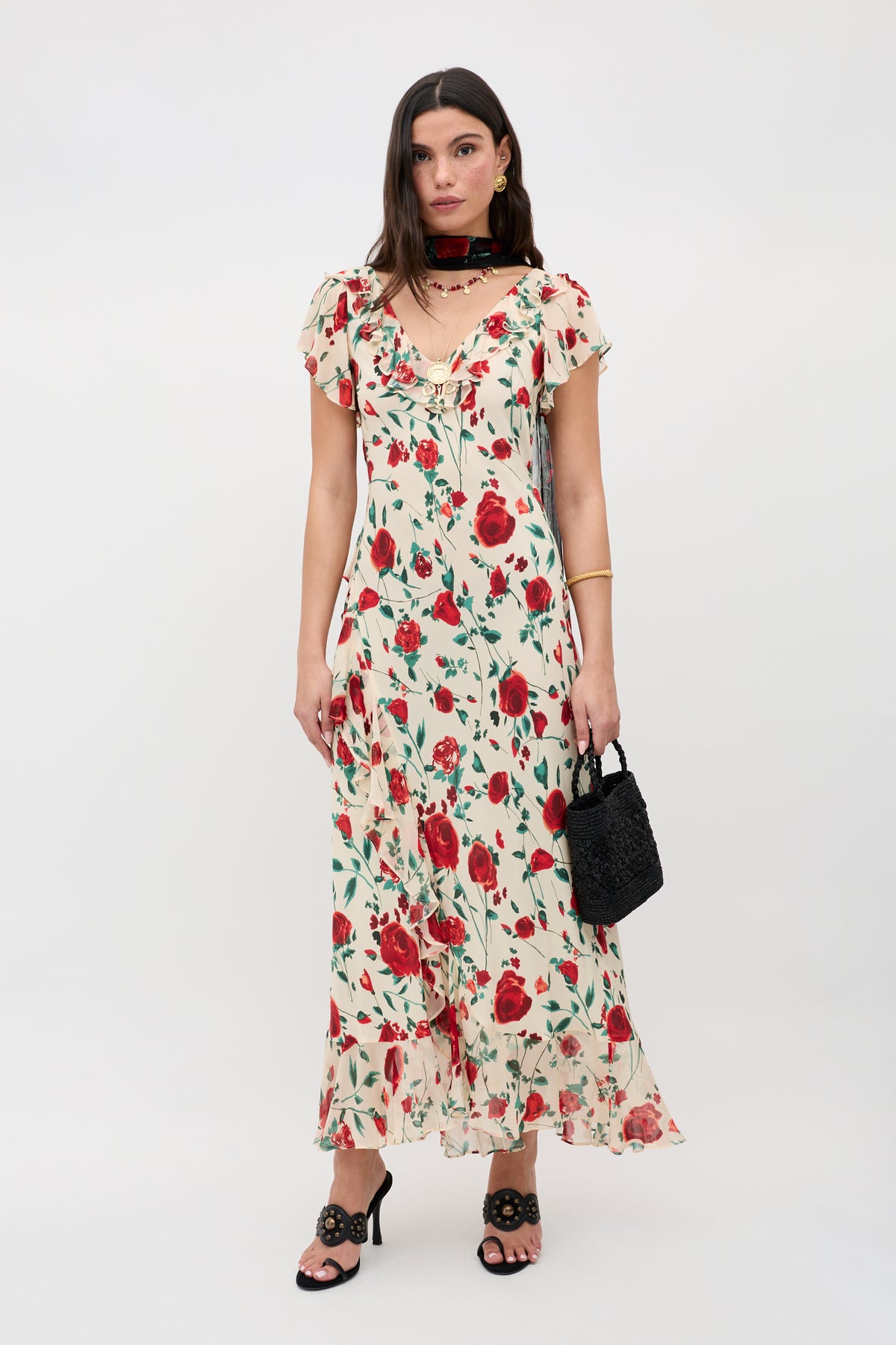 A woman stands against a plain background wearing the Meadow - Claudia Rose Cream floral dress with red roses, black heels, gold bangles, a dark choker, and holding a small black handbag.