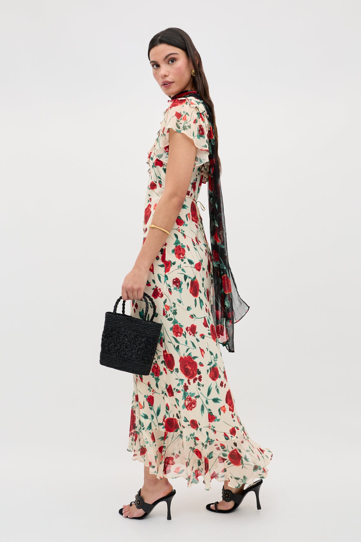 Woman stands sideways in the Meadow - Claudia Rose Cream ankle-length floral dress, featuring red and green patterns. She pairs it with black heeled sandals, a textured black handbag, and a sheer black scarf over her shoulders against a plain backdrop.