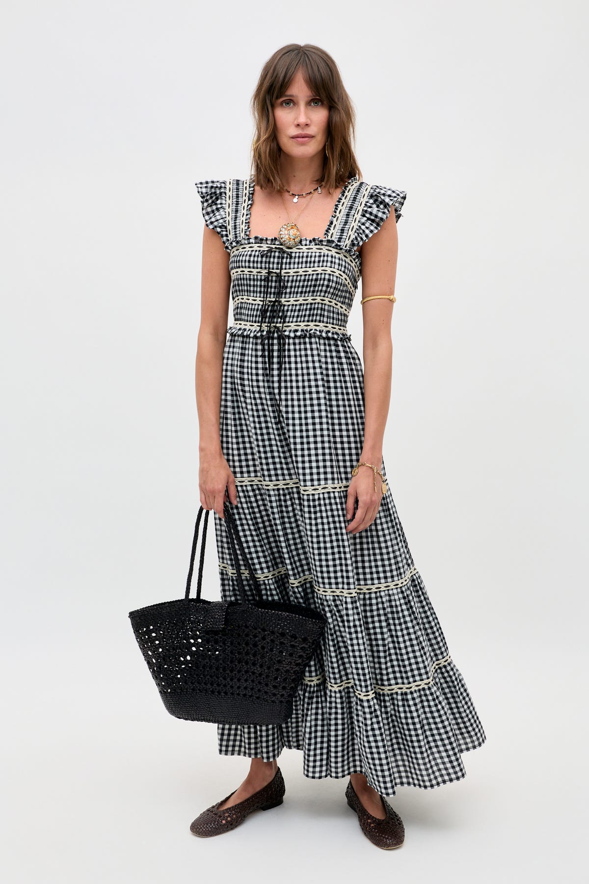 A woman stands against a plain background wearing the Chandra - Beth Check Black maxi dress with ruffled sleeves. She completes the look with a black woven tote, brown flats, layered necklaces, and an arm cuff.