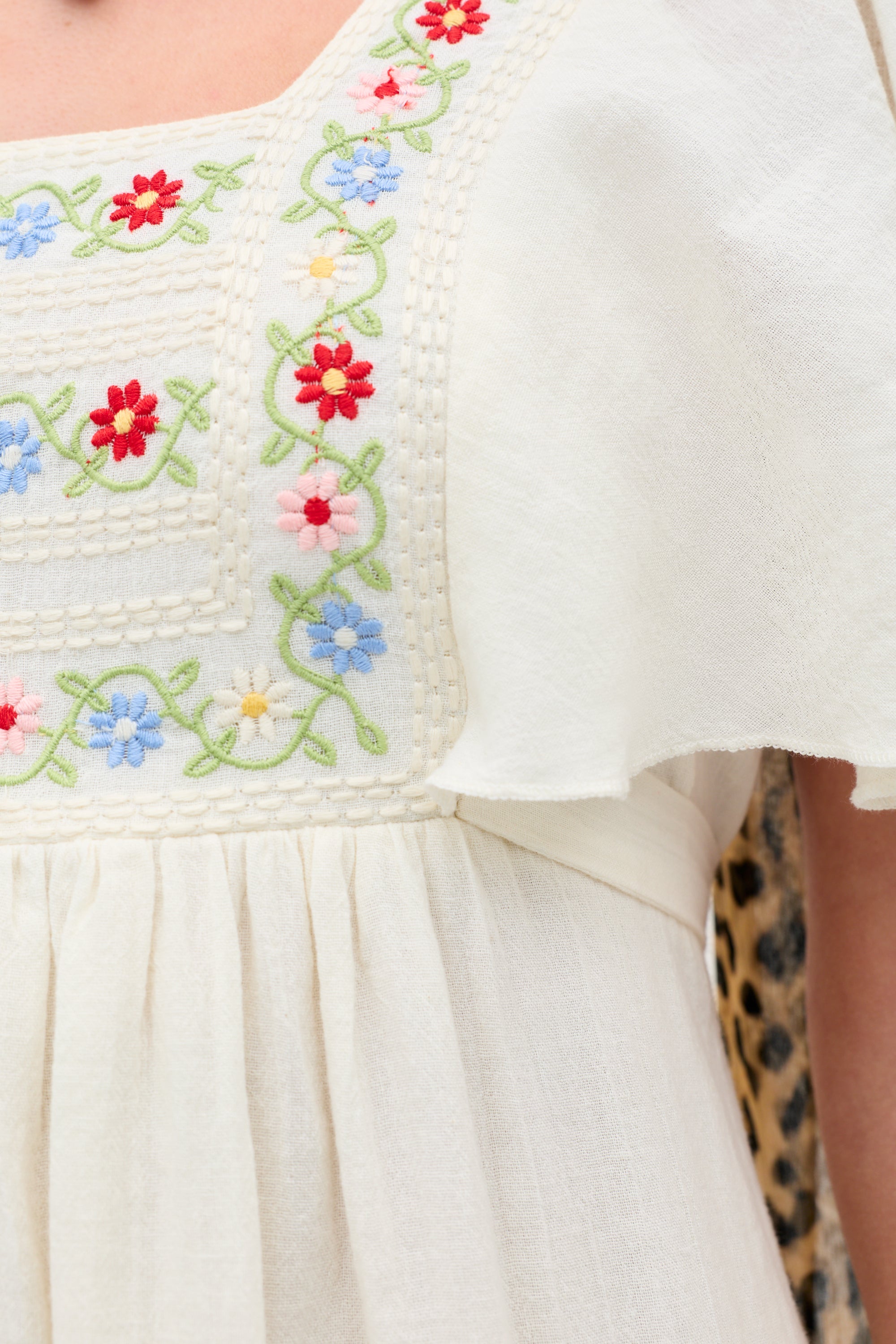 A close-up of the Jimena - Genoa Embroidery Cream dress, featuring colorful floral embroidery in red, pink, blue, and green on soft cream fabric. The dress has a square neckline and a gentle, flowing silhouette.