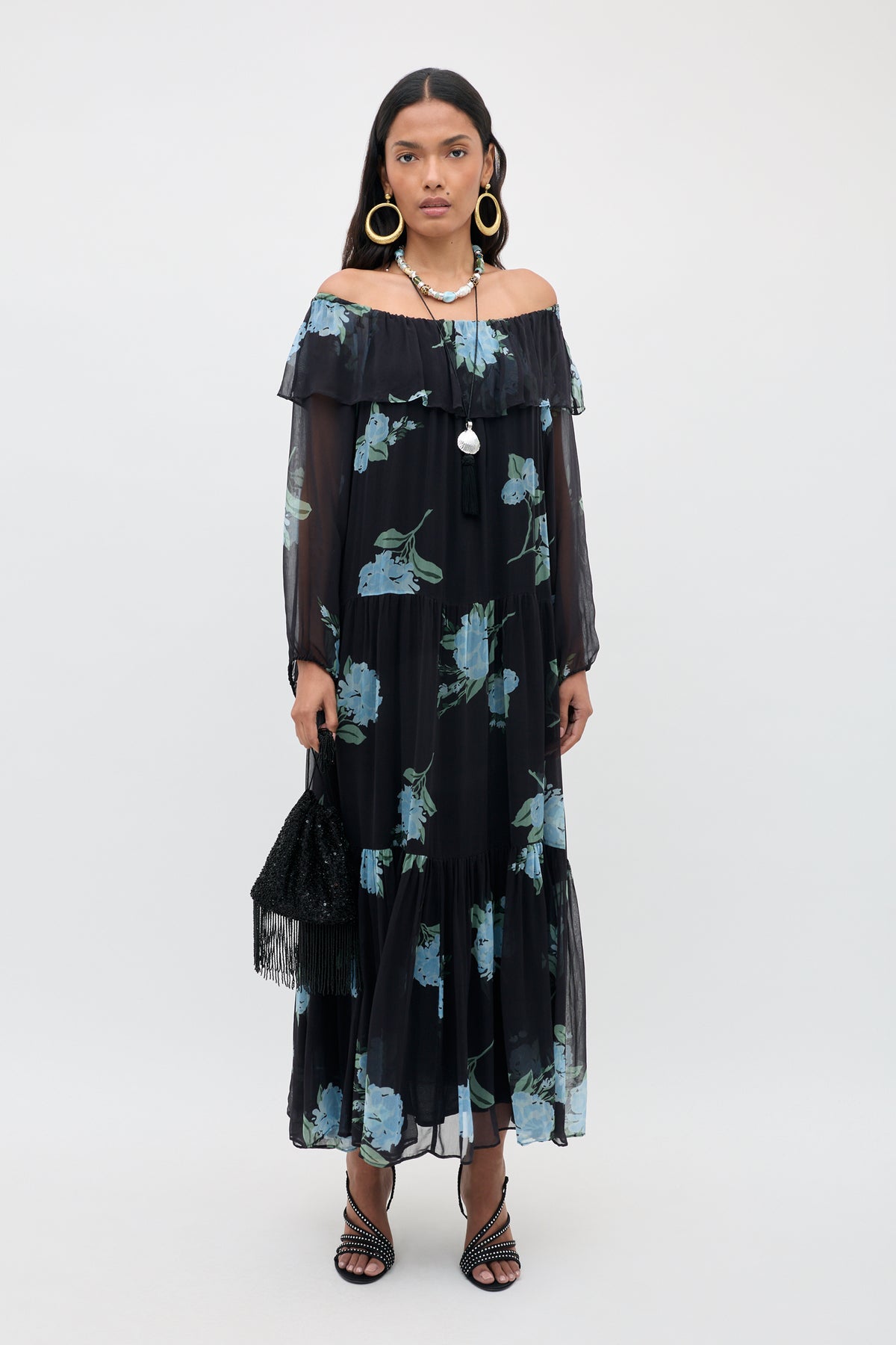 A woman models the Lela - Peonies Scatter Charcoal dress, featuring a black off-the-shoulder silhouette with blue floral print and sheer long sleeves, paired with large hoop earrings, layered necklaces, a black fringe handbag, and strappy black heels against a plain light background.