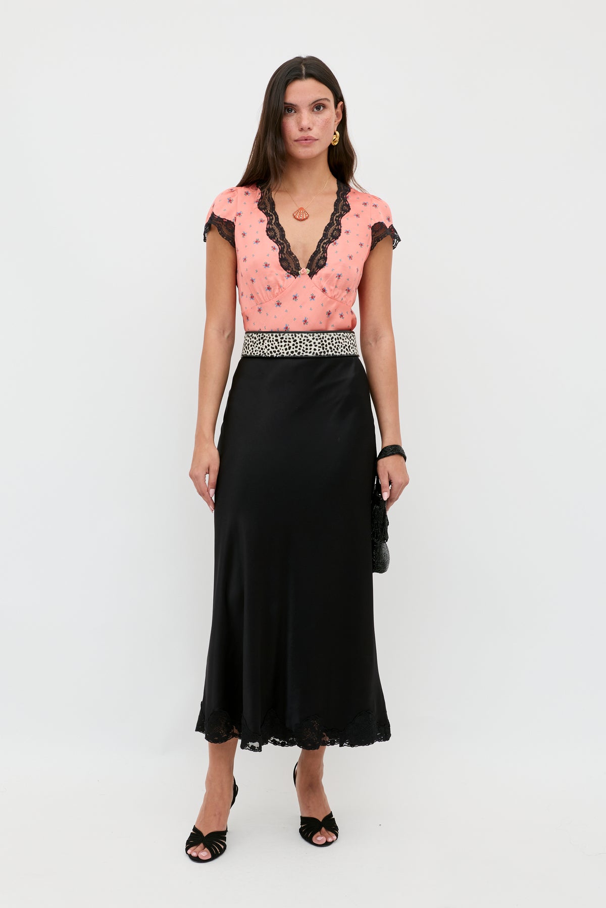 A woman stands against a white background wearing the Crystal - Black satin midi skirt with lace hem, a peach short-sleeved top with black lace trim, black heels, and holding a small black handbag.
