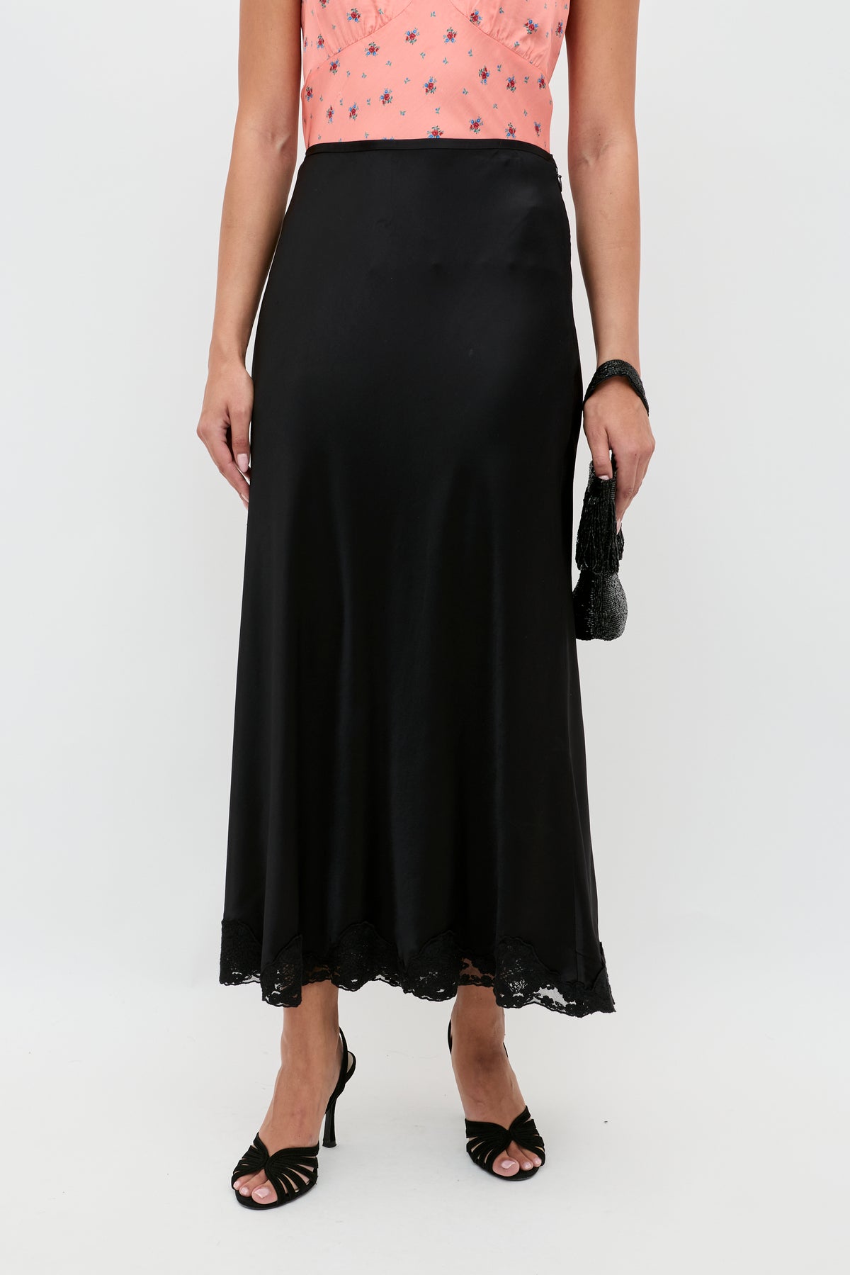 Wearing a peach sleeveless top and a black Crystal - Black satin midi skirt with lace trim, holding a black clutch and wearing black high heel sandals. Only the lower body is visible.