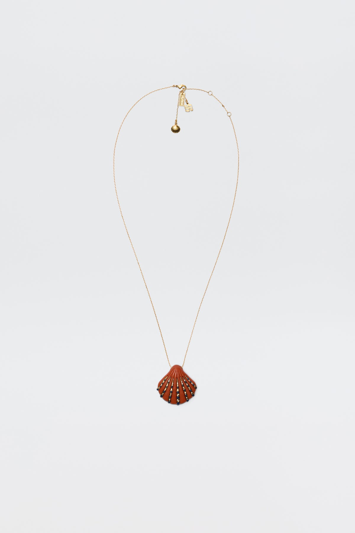 The Leanira - Orange necklace features a scallop shell-shaped pendant with small decorative studs on a gold-plated chain, shown against a plain white background.