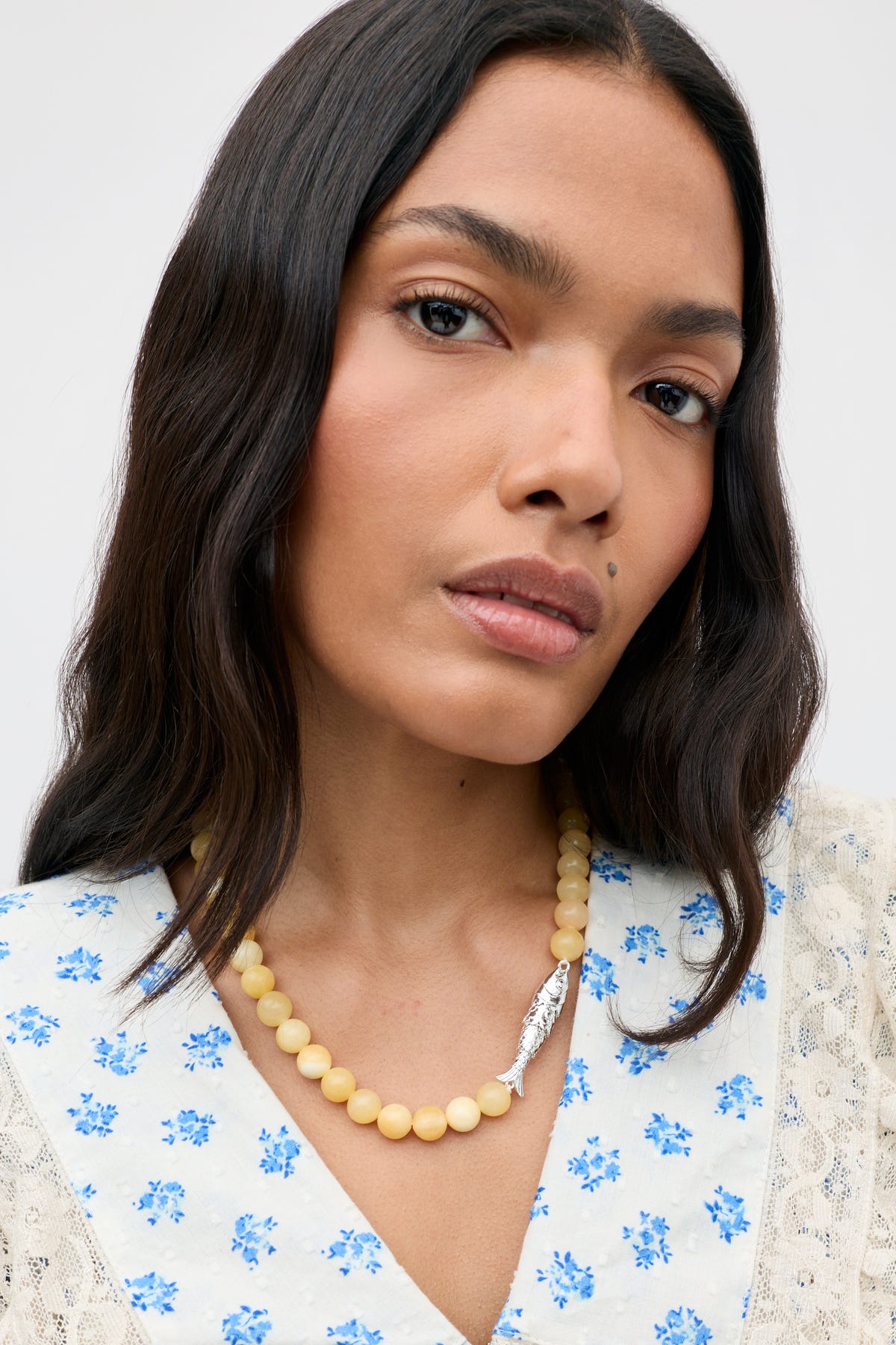 A woman with long dark hair wears the Amoura - Lemon cream beaded necklace featuring a silver feather pendant, paired with a white blouse adorned with blue floral prints. She looks directly at the camera against a plain background.