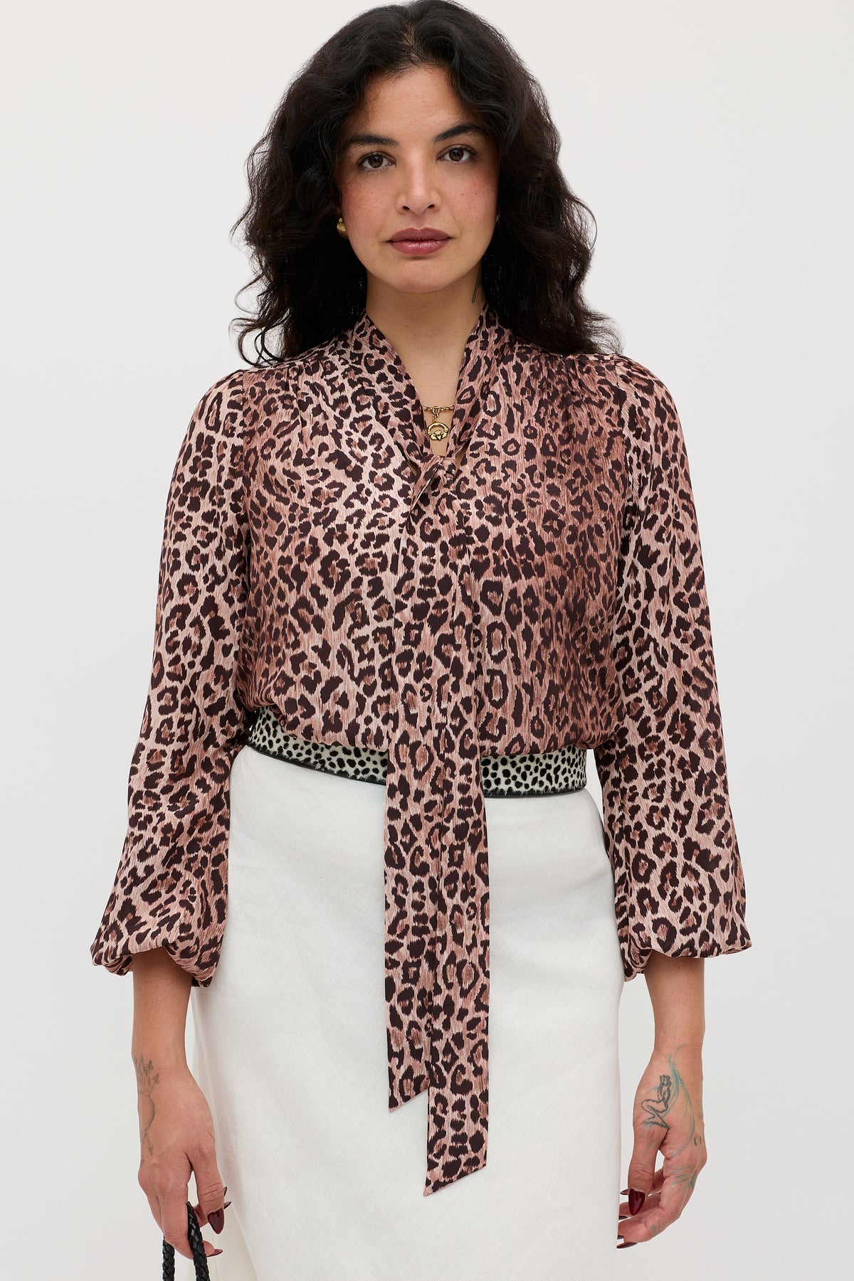 A woman with wavy dark hair wears the Moss - Leopard Brown blouse, a vintage-inspired pink and brown leopard-print silk top with a long necktie, paired with a white skirt and patterned belt, against a plain white background.
