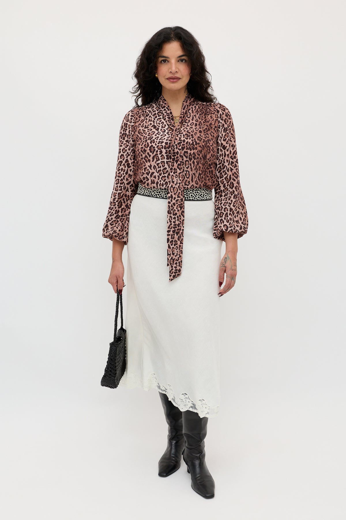 A woman stands against a white background wearing Moss - Leopard Brown, a vintage-inspired silk blouse, paired with a white lace-trimmed midi skirt, black knee-high boots, and holding a black woven bag.