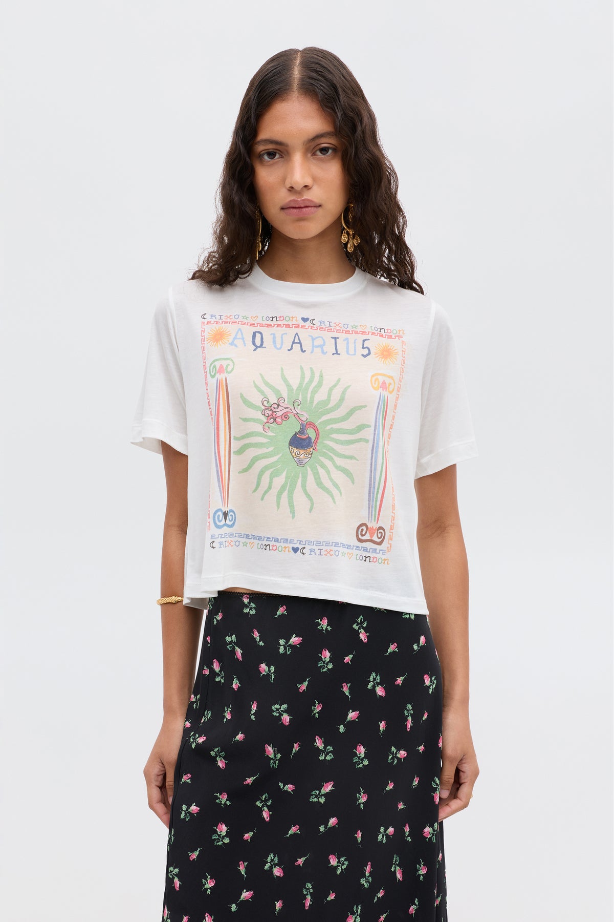 A woman with wavy dark hair wears the Ria - Aquarius white graphic T-shirt featuring an Aquarius illustration and a black skirt with a pink floral pattern, standing against a plain light background.